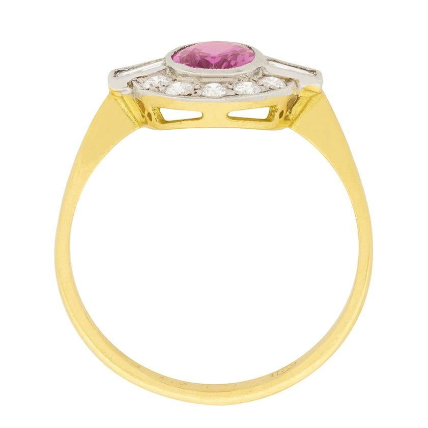 A 0.75 carat pink sapphire is encircled by a halo of diamonds. The oval cut pink sapphire has a beautiful vibrant colour. To either side of the sapphire are a baguette cut diamond, each is 0.10 carat.  Ten round brilliant diamonds form the halo and
