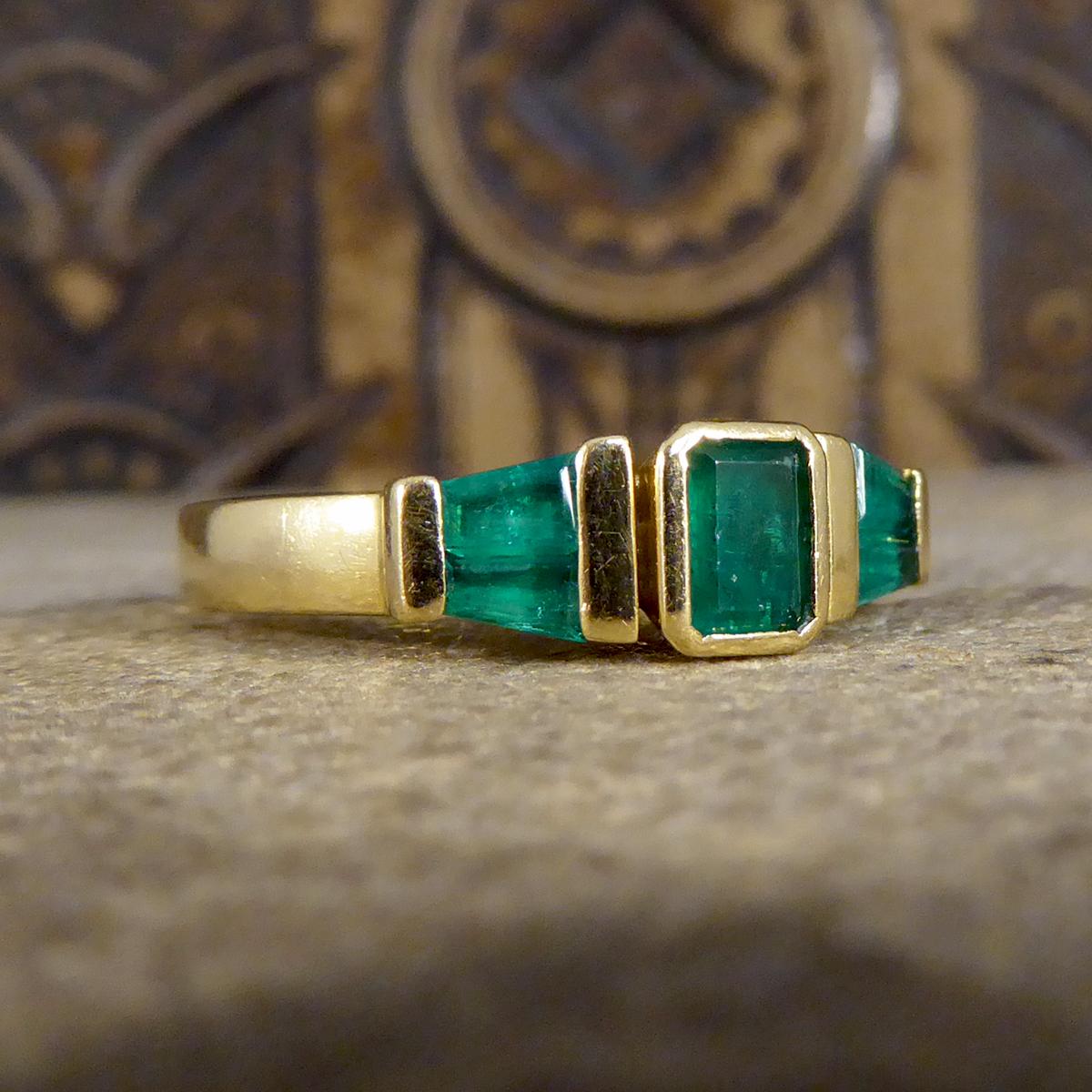 This ring features five Emeralds with the centre Emerald being Emerald Cut and two Tapered Baguettes on either side of the centre weighing approximately 1.00ct in total. There centre Emerald is set in a rub over collar setting and the side shoulder