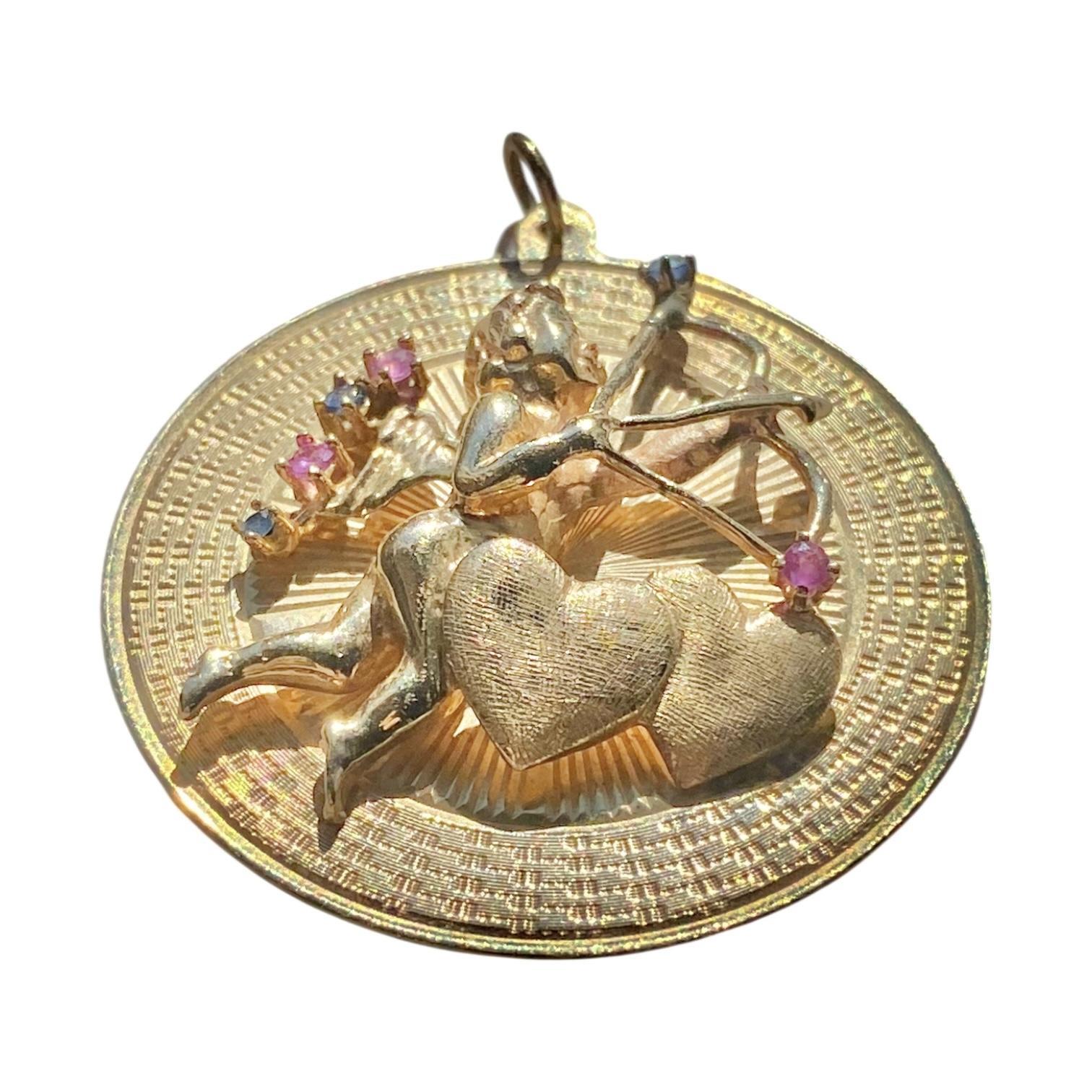 Réalisé en or jaune 10 carats, le pendentif présente un cupidon avec un arc et une flèche, accentué par des motifs en forme de cœur. La pièce est ornée de rubis et de saphirs bleus taillés en rond.
3 Rubis : 0,10ctw.
3 saphirs bleus :