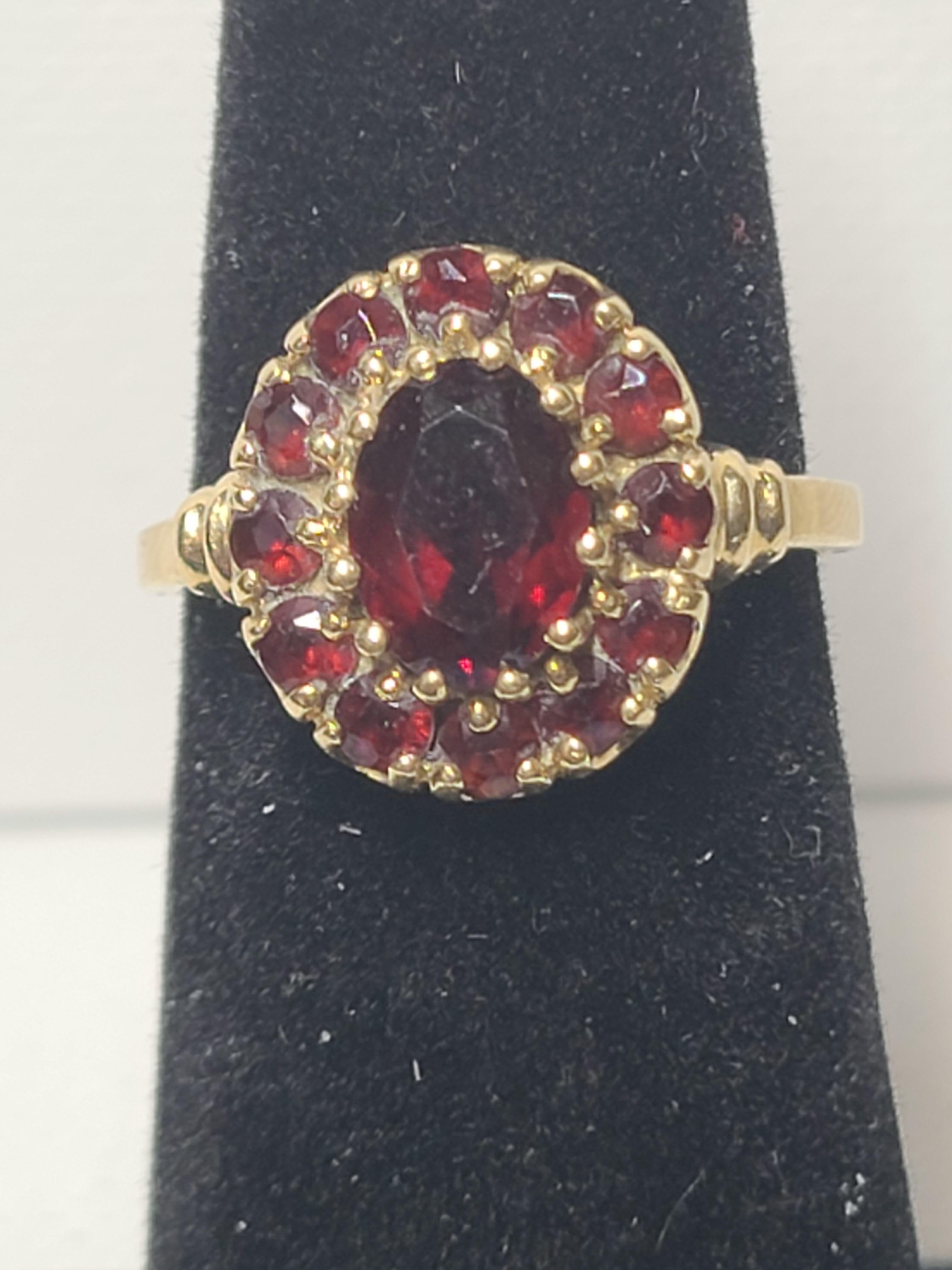 Dieser Vintage-Cocktailring aus einem Nachlass verfügt über einen 8x6 mm großen ovalen Mittelstein, der von einem Kranz aus 12 kleineren runden Granaten umgeben ist und einen klassischen Look bietet.
Die Steine sind in einem 10K Gelbgold Fassung,