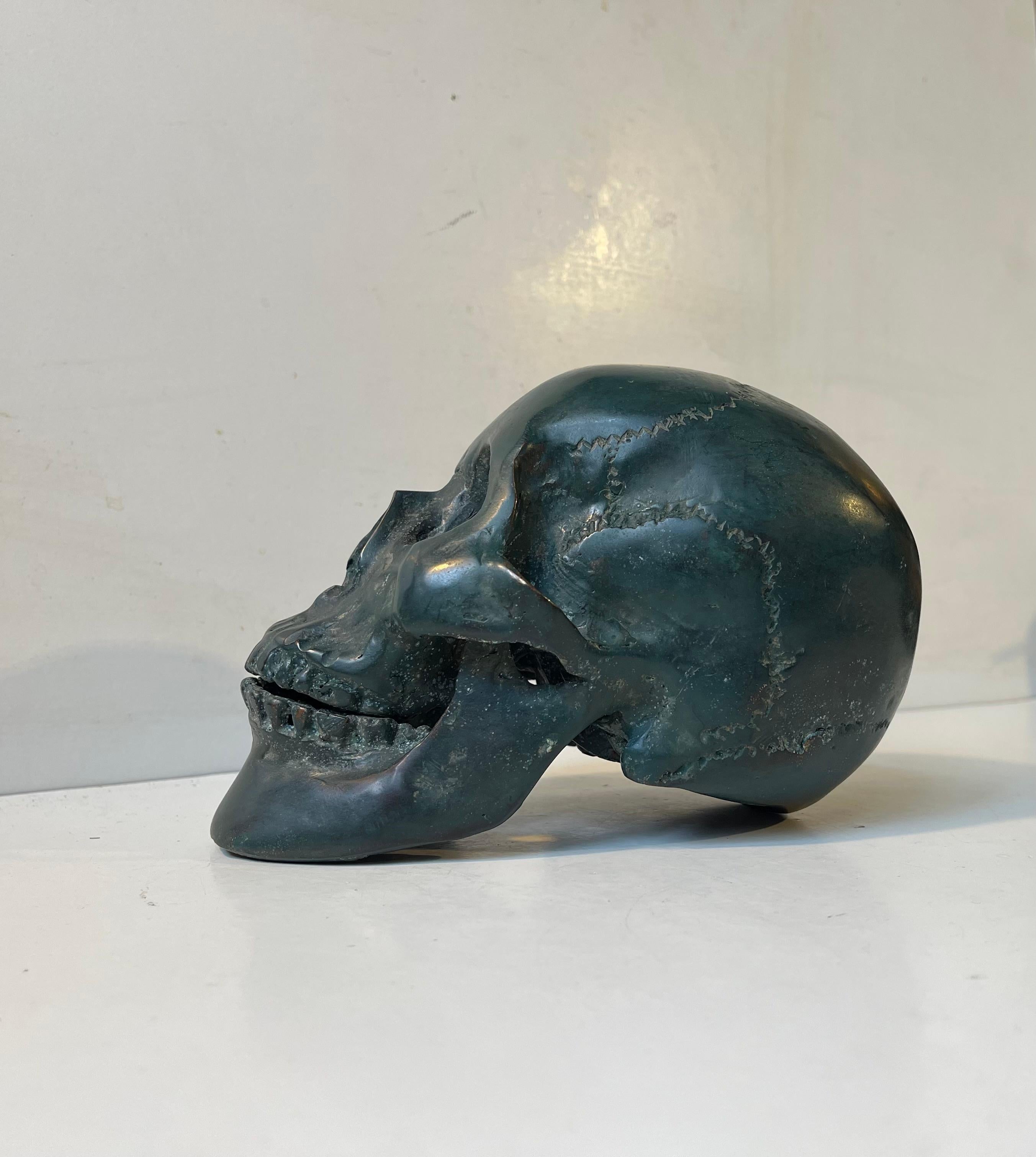 Modello in scala 1:1 di un teschio umano in bronzo verde verdastro (patina artificiale). Si tratta di un calco di un vero cranio umano, probabilmente femminile, completo di denti, mascella inferiore e superiore. Questo particolare esempio ha