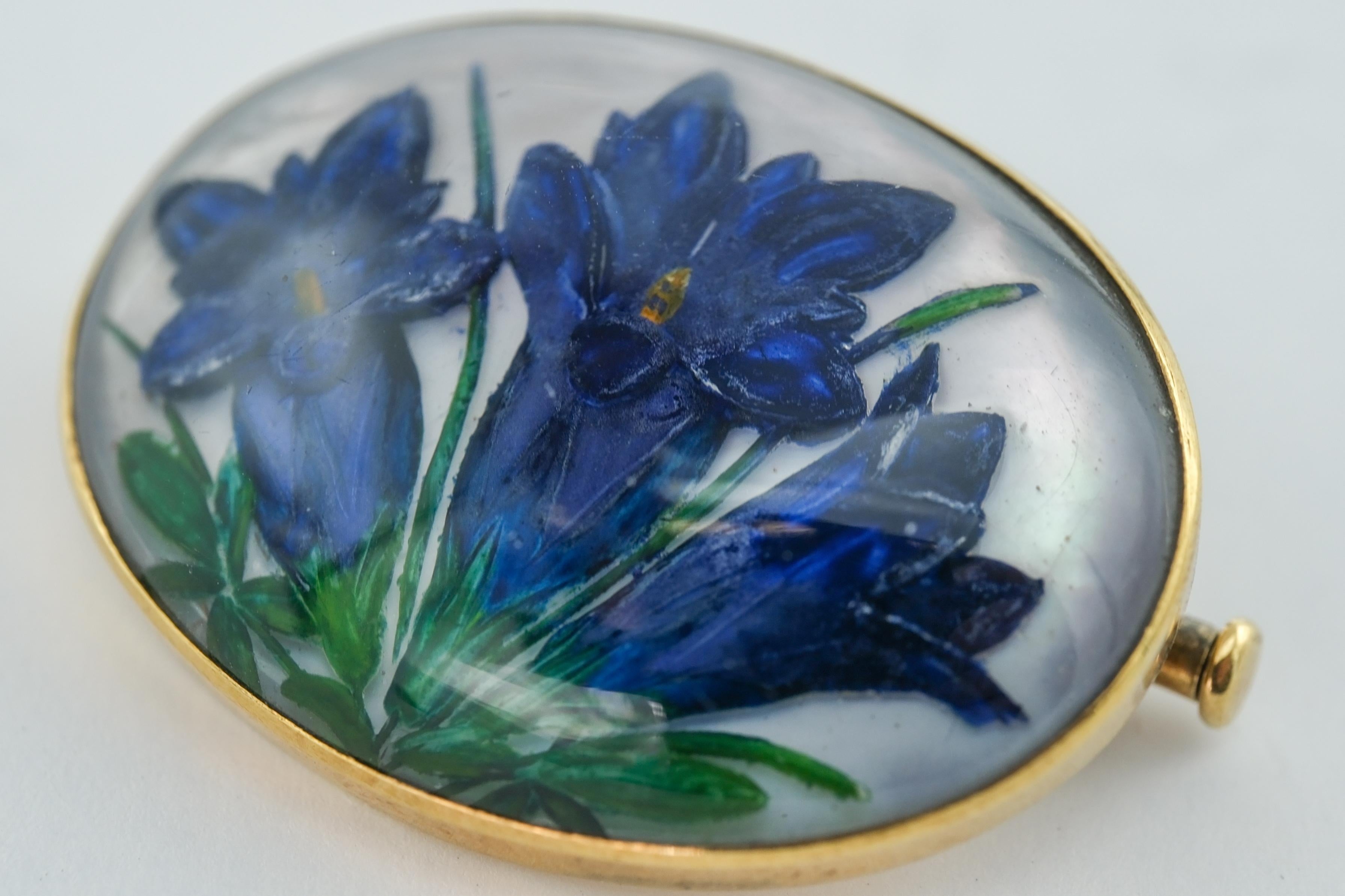 This 14 karat yellow gold pin showcases a masterfully executed reverse painting intaglio of three blueish-purple flowers. Reverse painting intaglio is a specialized technique wherein a design is painted in reverse on the underside of a transparent