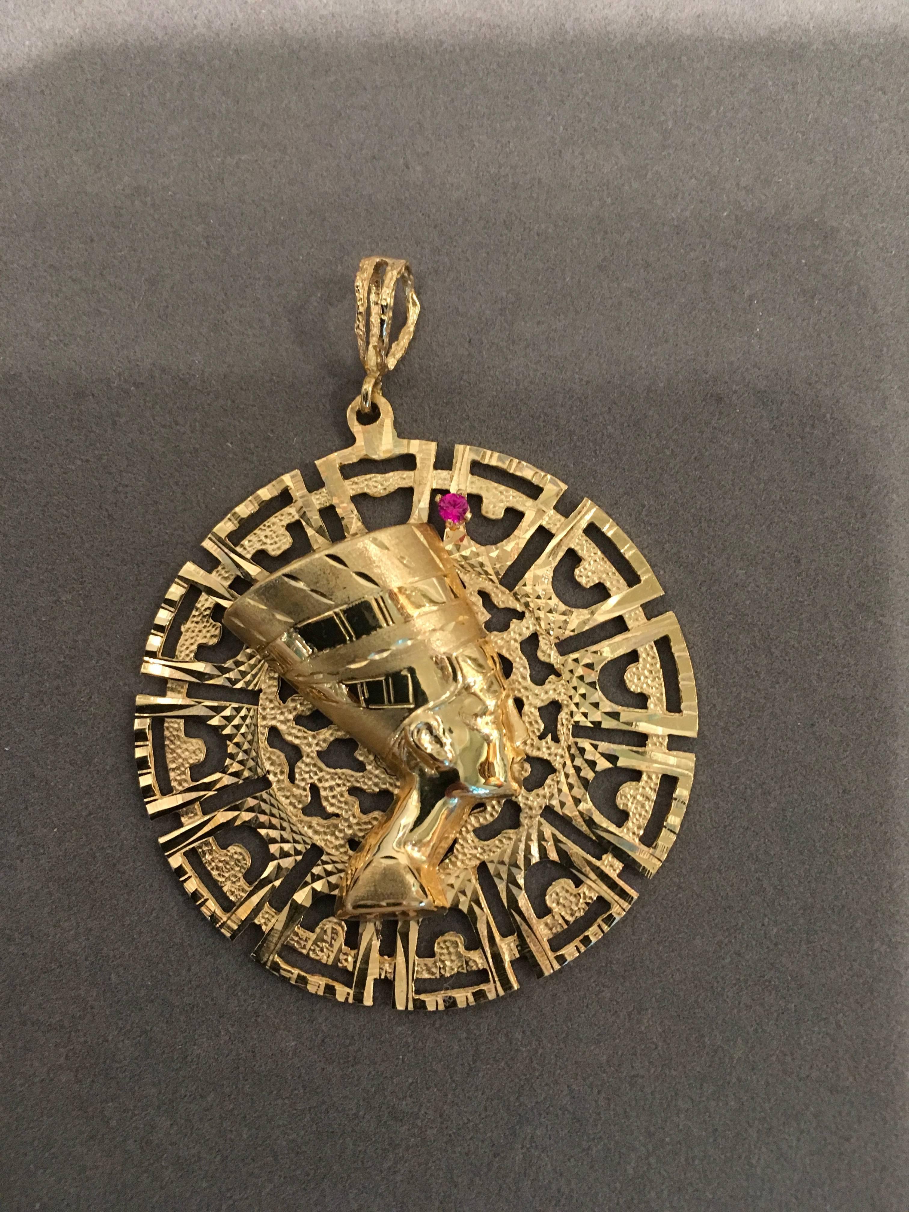 A vintage pendant featuring a bust of Egyptian Queen Nefertiti over a geometric background in 14K gold, circa 1975. The pierced disk has a variety of textures and cut-outs. Hollow profile of Nefertiti is mounted at the center with a bright pink