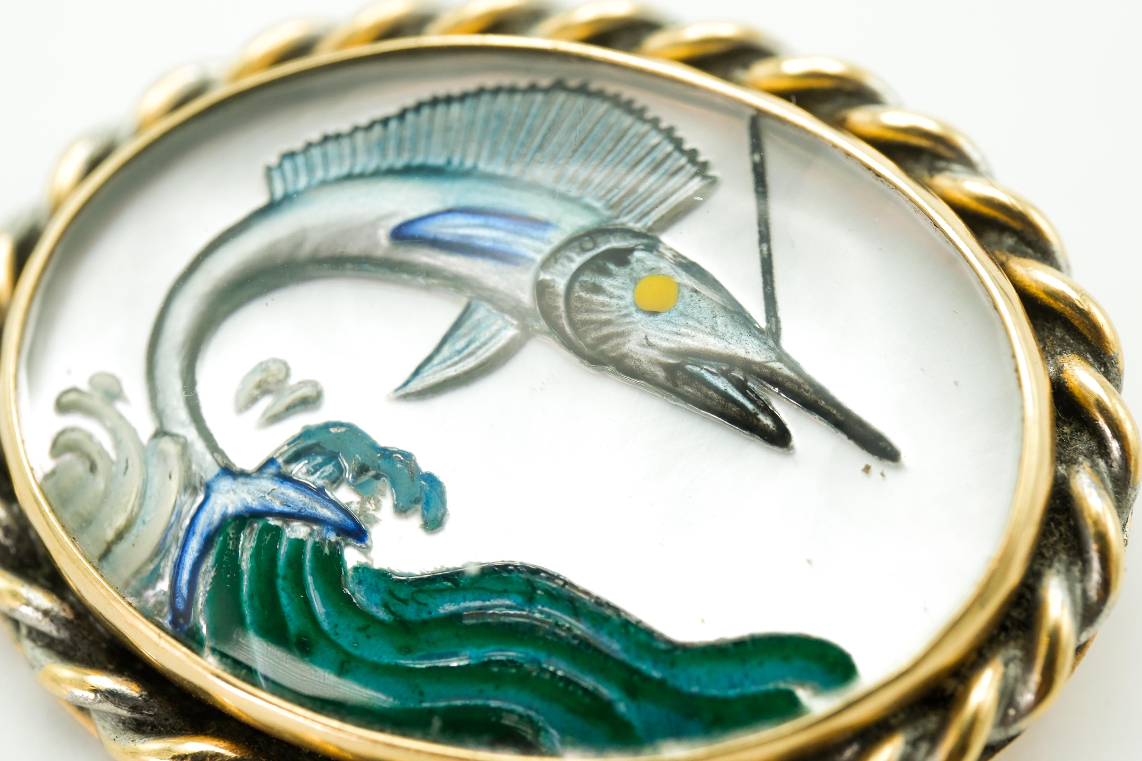 This vintage brooch is crafted from 14 karat yellow gold and features a reverse painting intaglio of a marlin. The depicted scene showcases an active marlin leaping dynamically from the ocean, captured intricately within the crystal. Framing the