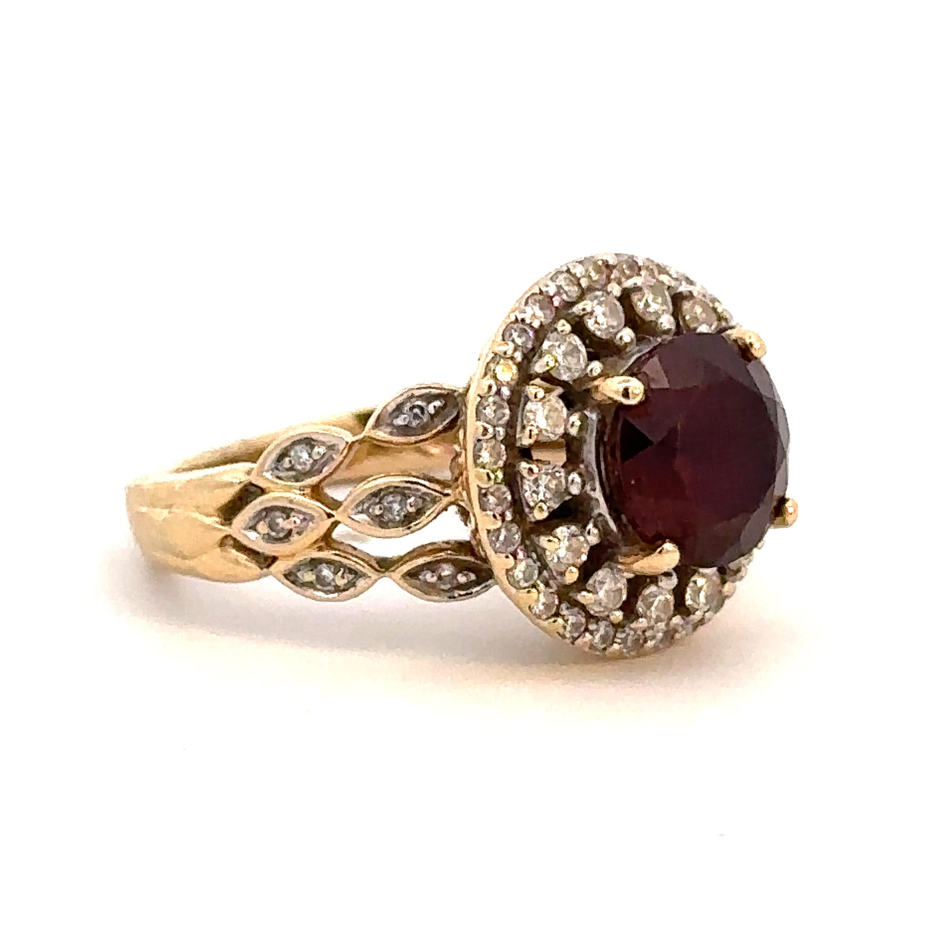 An ornate vintage 14k yellow gold ring presenting a garnet within a double halo of diamonds, with additional diamonds along the shank. The garnet has a rich red hue and weighs approximately 2.43 carats. The ring is a size 5.5, but can easily be