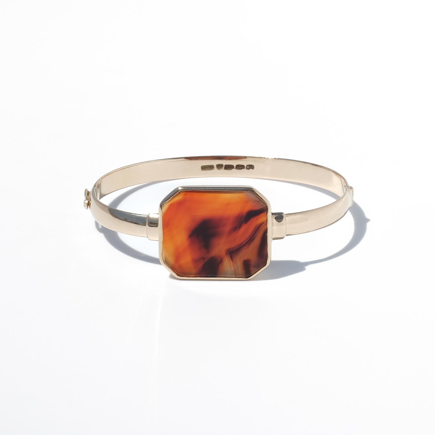 This fixed bracelet is crafted in 14 karat gold. The band is smooth, slightly rounded, and solid, forming a continuous oval shape with a hinged opening. At the center sits an octagonal agate, bezel-set in a clean, polished frame. The agate shows