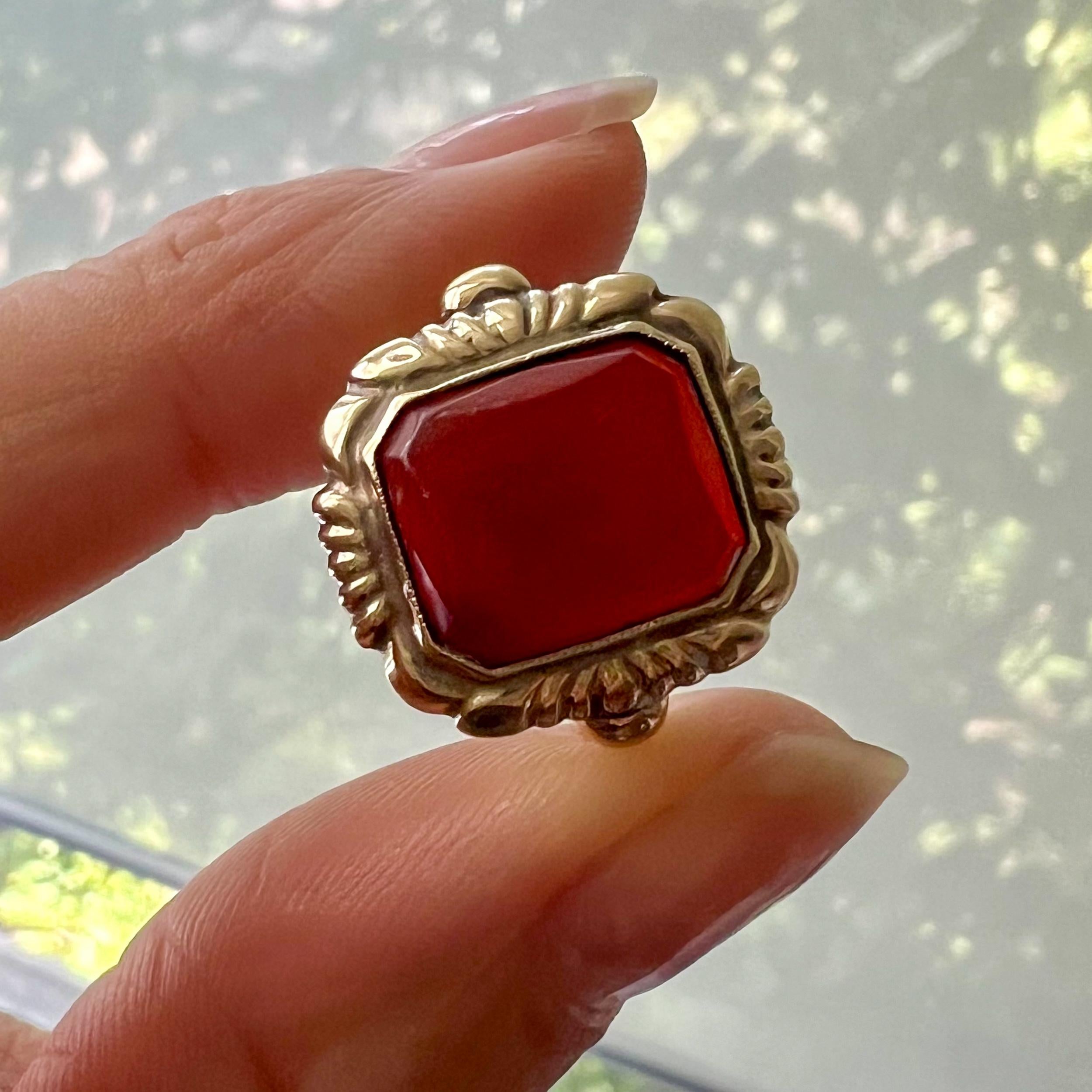 Ein quadratischer Ring aus 14-karätigem Gold, besetzt mit einem dunklen Karneol-Edelstein. Die Goldfassung dieses Rings ist an der Vorderseite und an den Schultern mit einem Rillenmuster versehen. Der Karneol hat eine schöne dunkelrote Farbe, ist