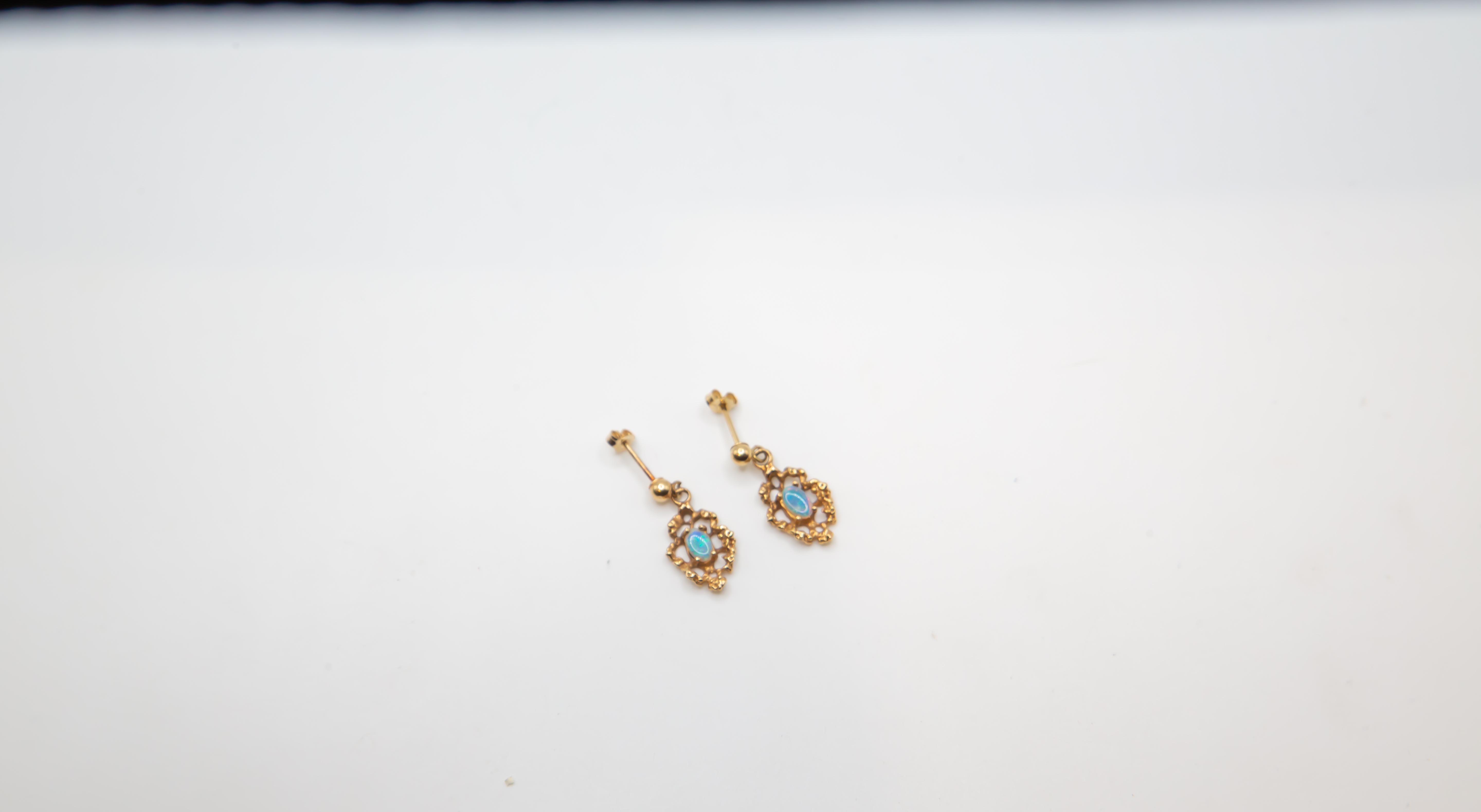 Un affascinante paio di orecchini vintage in oro giallo 14K con opali naturali luminosi incastonati in cornici a volute. Ogni orecchino sospende un'opale ovale incastonata in un castone che brilla con l'accattivante iridescenza blu, verde e rosa