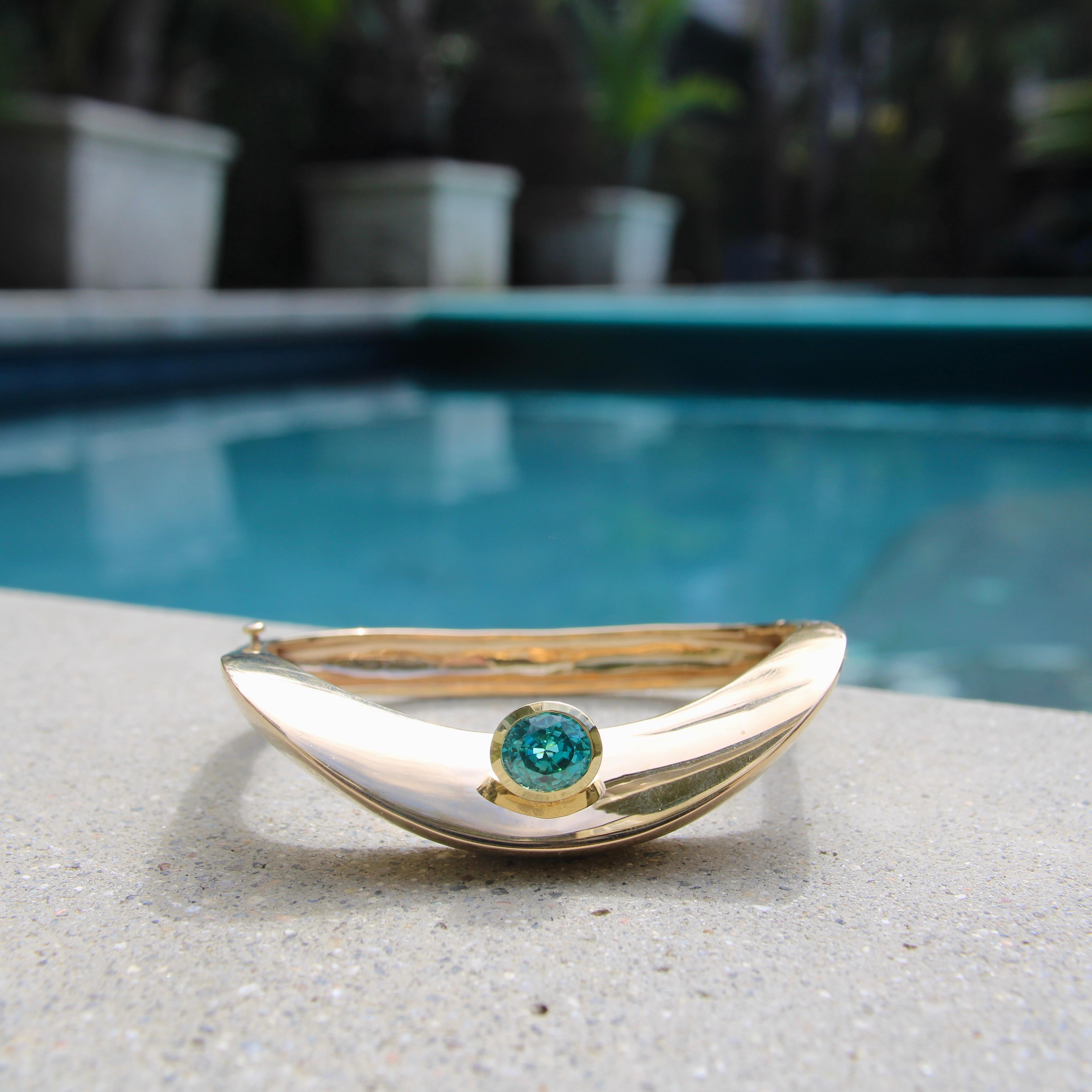 Circa the 1970’s, this 14k gold modern bracelet has a wave-like shape with a knife edge along the top side. The swoop of the knife edge makes the eye follow the piece to a bezel set blue zircon nestled in the center, perfectly complimenting the