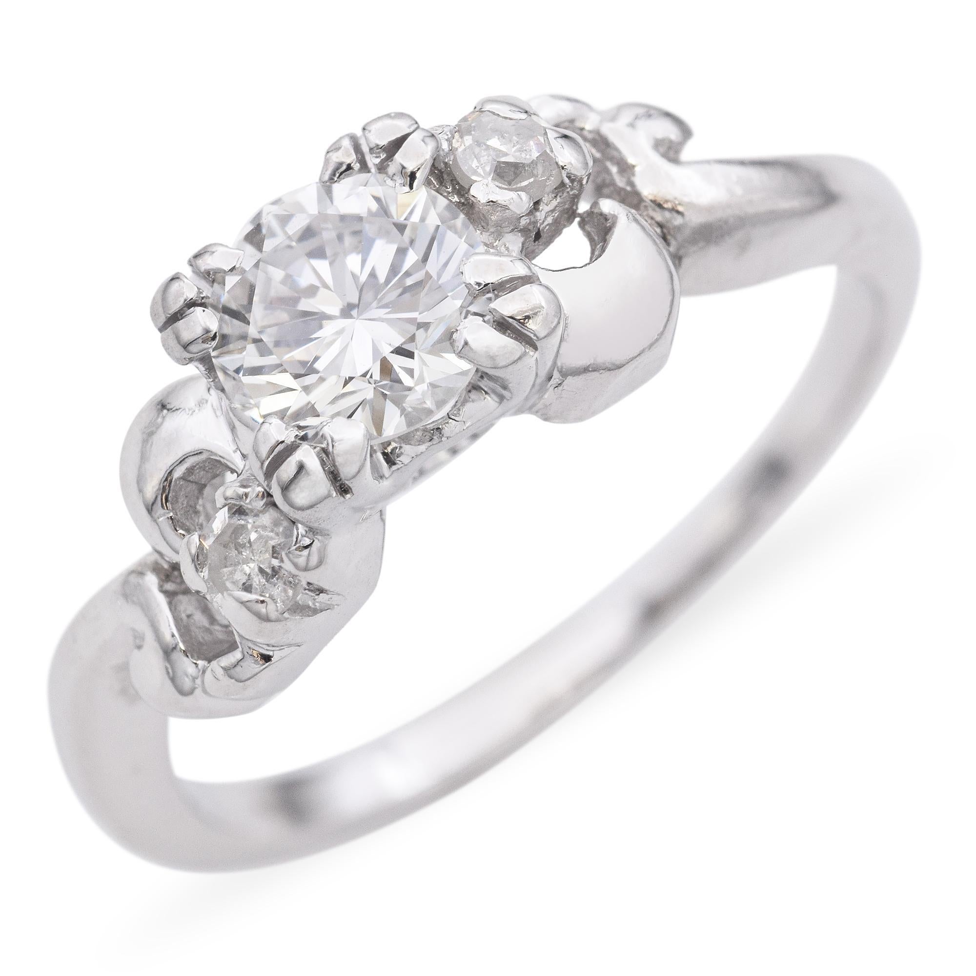 Questo affascinante anello presenta un diamante rotondo centrale con taglio a brillante, affiancato con eleganza da due diamanti più piccoli. L'intricata montatura presenta un design unico e fluente che esalta la brillantezza delle pietre.