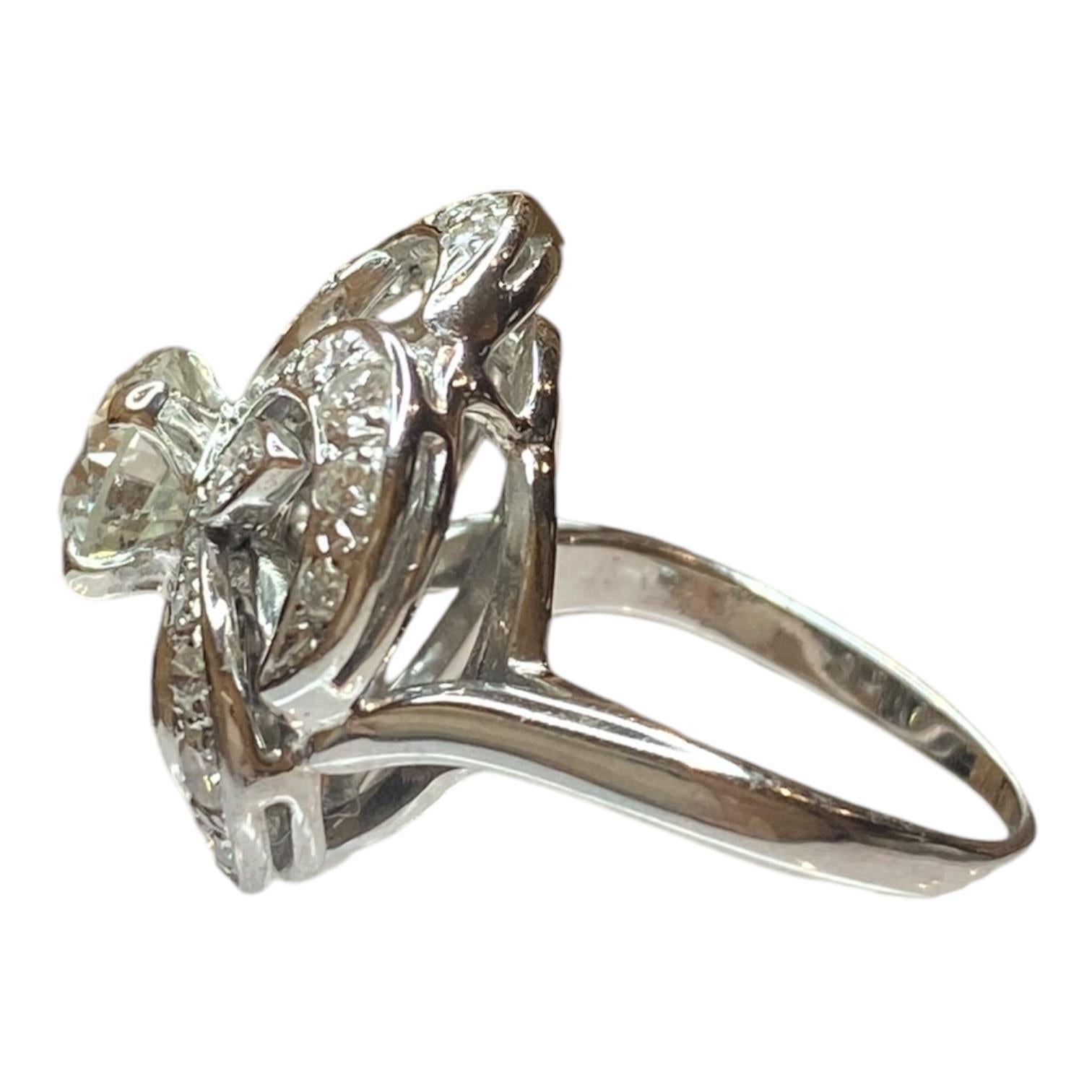 Anillo vintage de oro blanco de 14 quilates con un intrincado motivo floral de pétalos calados y arremolinados. El diamante central está engastado de forma prominente, rodeado de diamantes adicionales que realzan su brillo.
Diamante central: 1,17