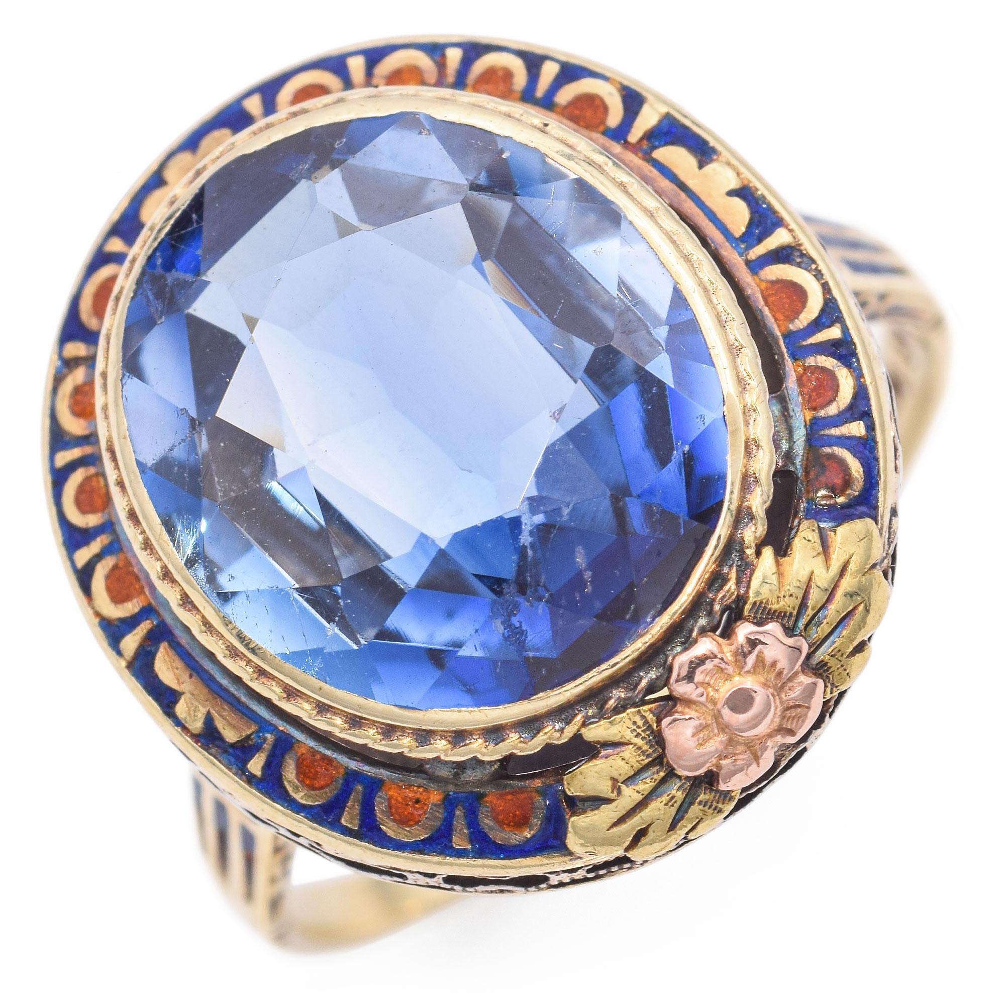 An exquisite ring featuring a large oval blue lab grown sapphire, set in a detailed gold band. The band is adorned with intricate enamel work in blue and orange hues, accented by floral motifs. Crafted with precision, this piece showcases a