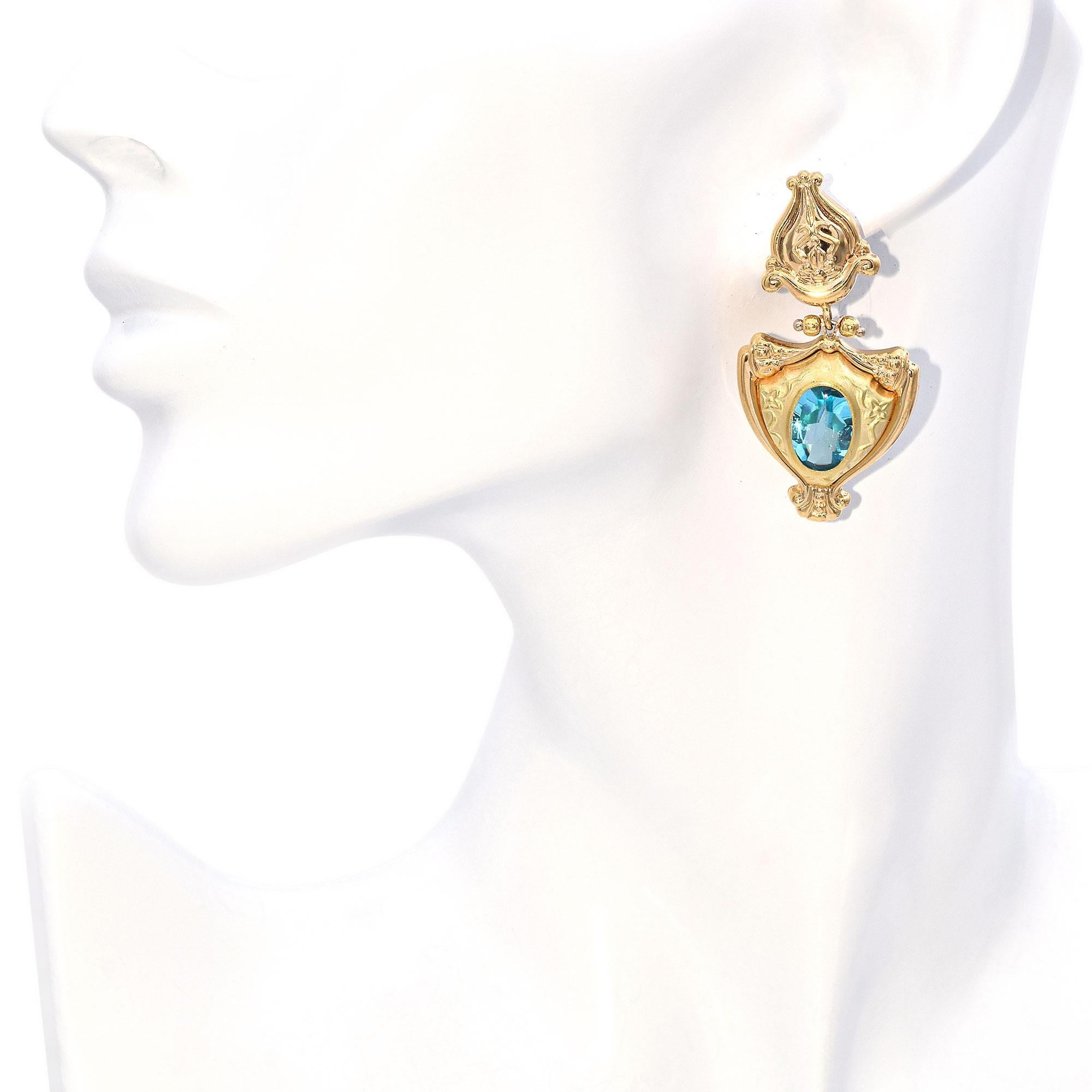 These exquisite earrings feature an intricate design with ornate gold detailing. The top section showcases a floral motif with elegant curves. Below, a larger section is adorned with a central oval blue gemstone, set within a decorative gold frame.