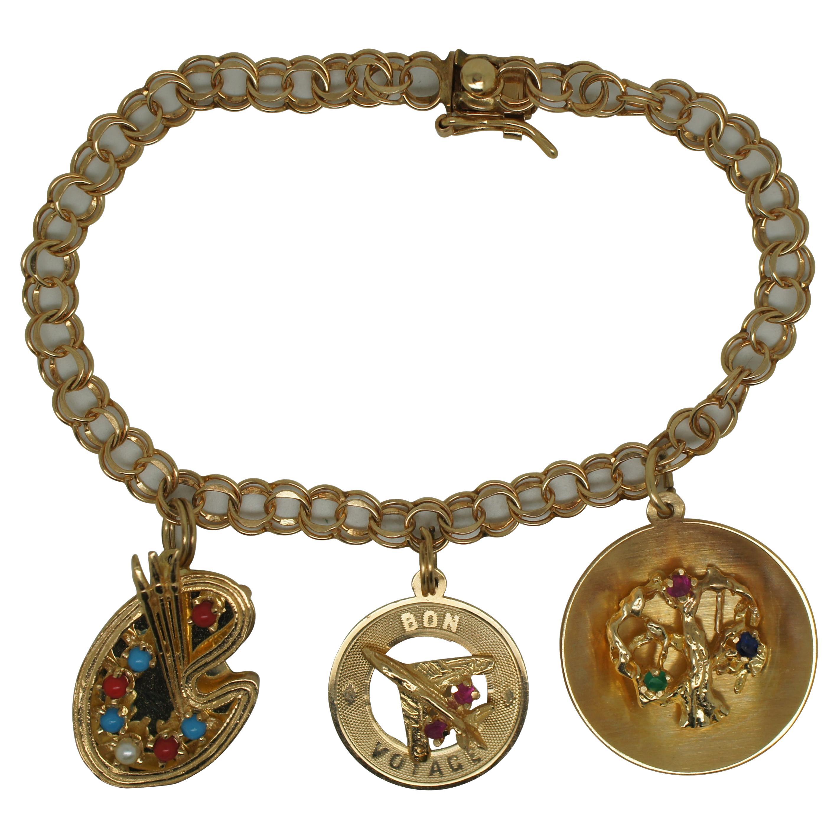 Vintage 14k yellow gold double link charm bracelet with open box clasp. The gold and jeweled charms include a paint palette with coral, turquoise and a pearl, a tree of life with emerald, ruby and sapphire, and a Bon Voyage jet airplane / plane with