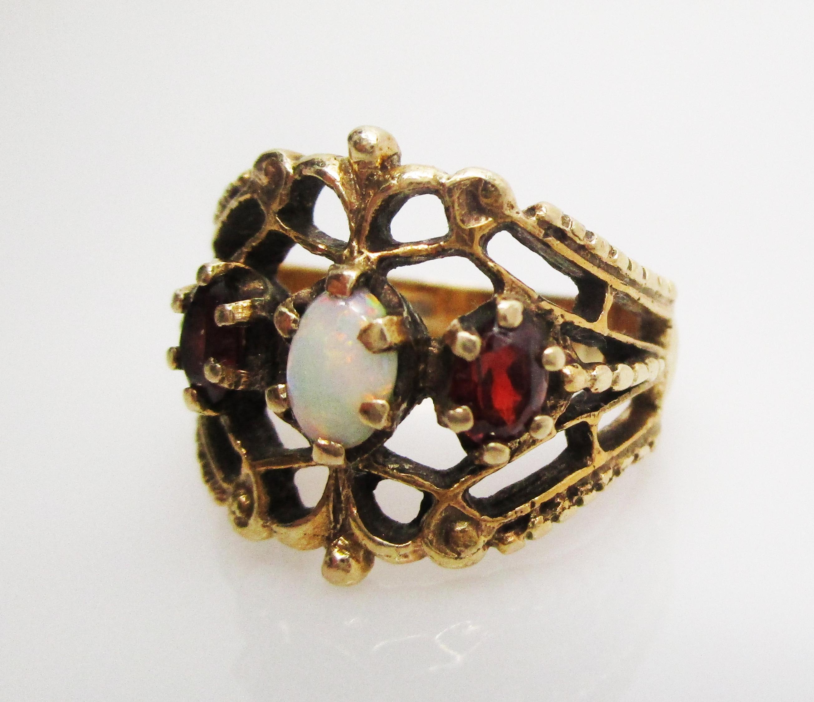 This is a fantastic 14k yellow gold ring featuring a stunning fiery white opal center stone flanked by two beautiful garnet side stones. The filigree design of the ring creates a unique silhouette that is reminiscent of a royal crown. The negative