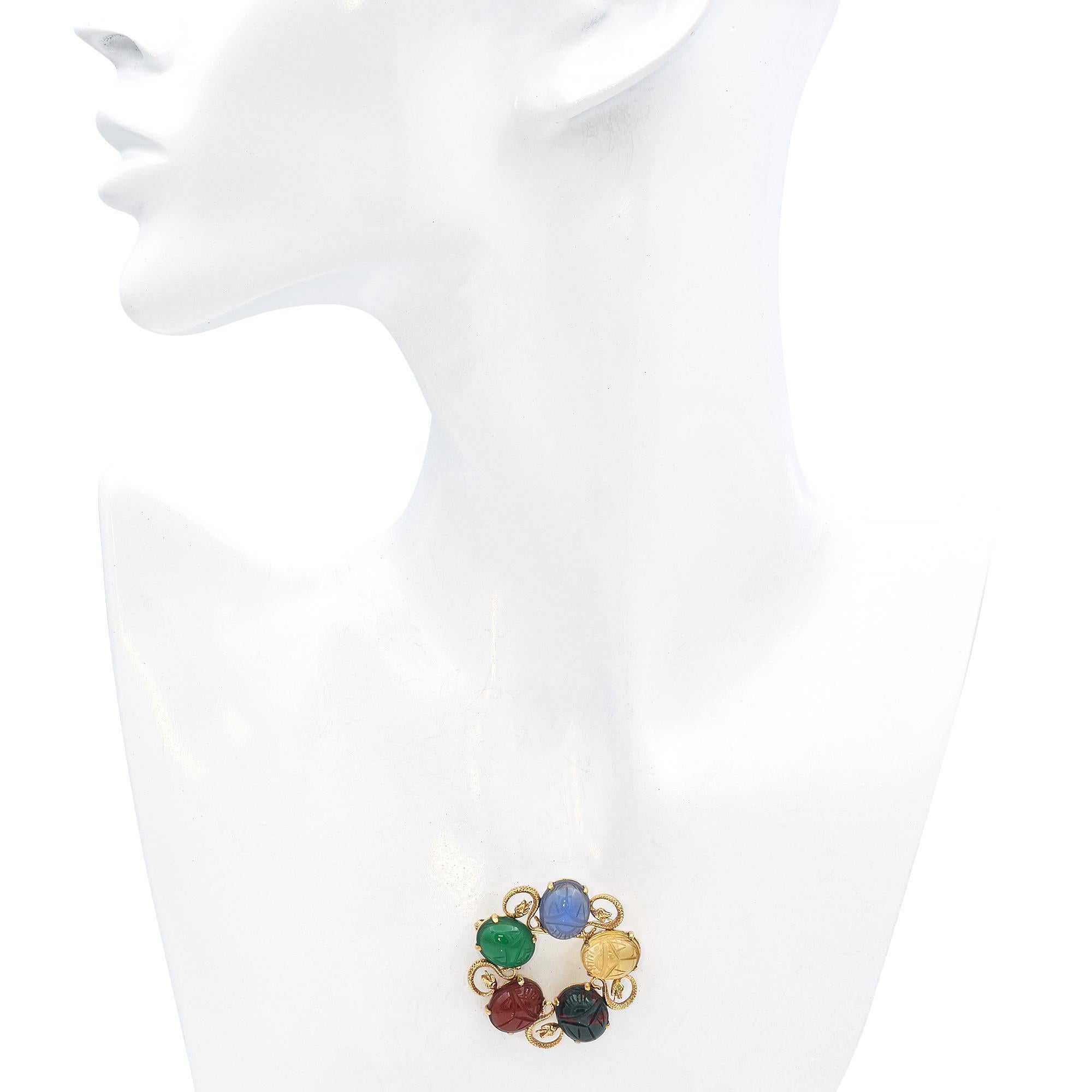 This exquisite brooch crafted in 14k gold features five intricately carved scarab gemstones in vibrant hues of blue, green, yellow, red, and dark green. Each scarab is securely set within a swirling gold framework adorned with detailed snake motifs.
