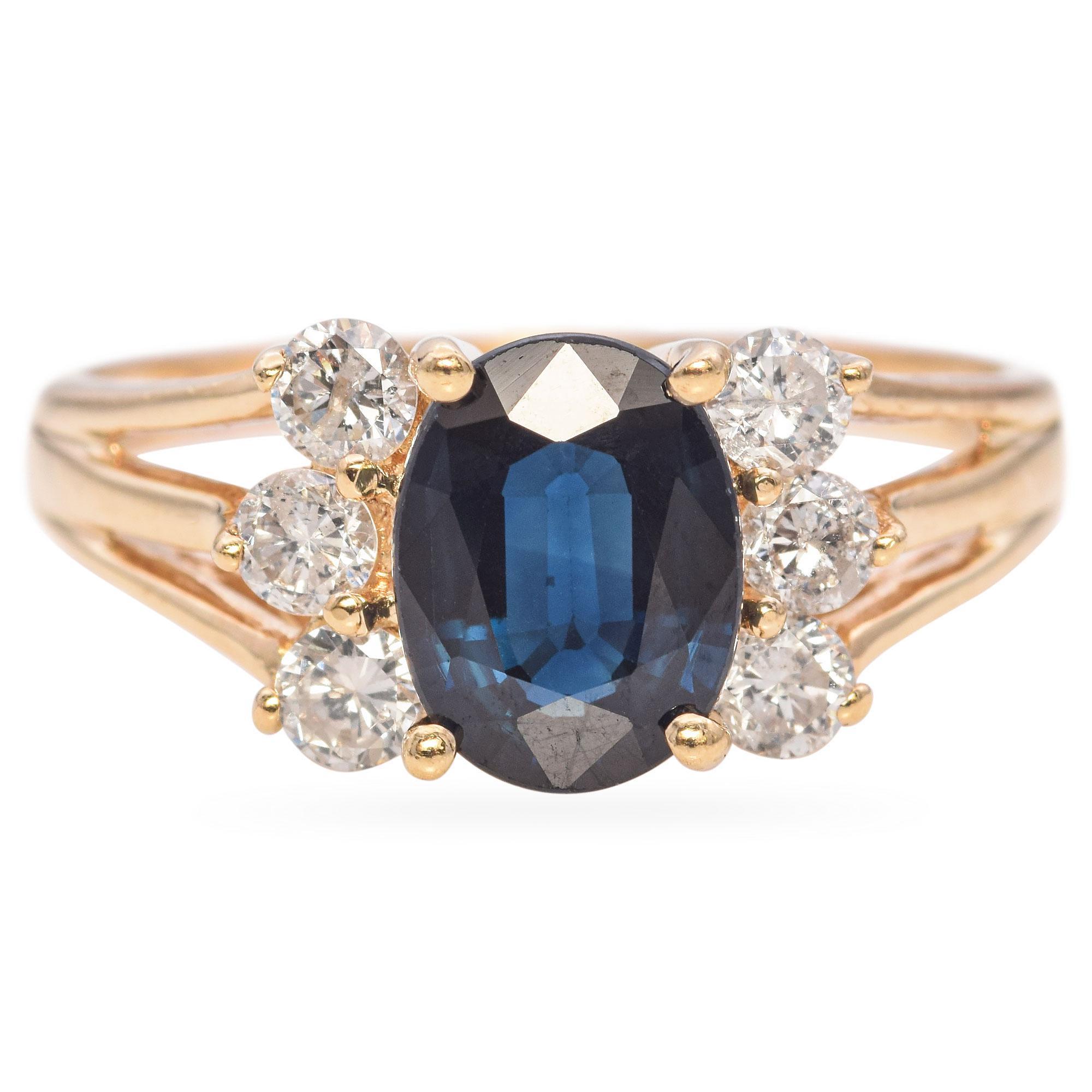 Bague élégante présentant en son centre un saphir bleu ovale saisissant, entouré d'une grappe de diamants ronds étincelants. Le saphir est solidement serti dans un anneau d'or chaud, qui se divise en un motif délicat, rehaussant l'élégance générale
