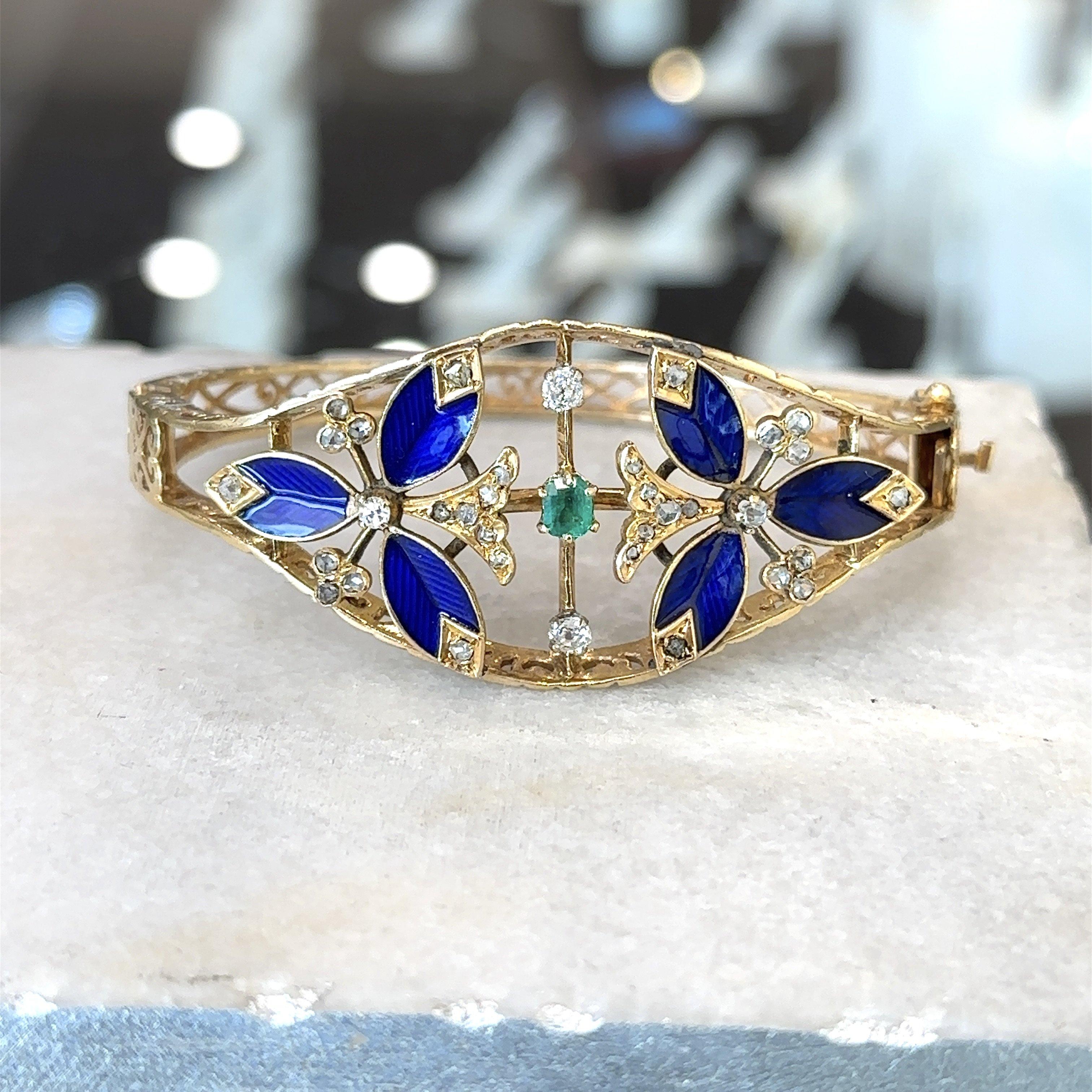 This beautiful hinged bangle bracelet dates from the 1950s. It is crafted from 14KT yellow gold with cobalt blue guilloche detailing and features approximately .70CT high crown rose-cut diamonds and one .35CT oval emerald. The decorative center of