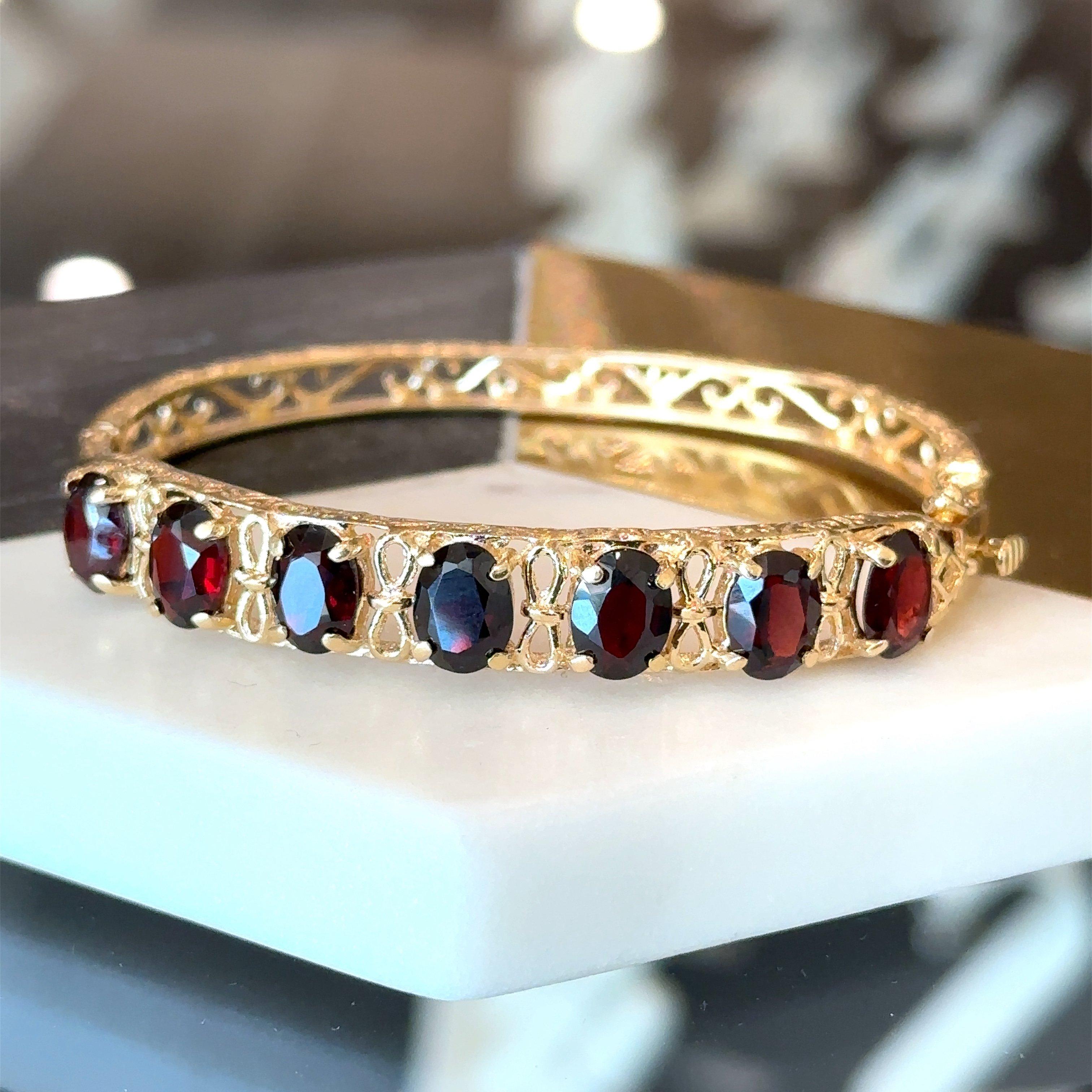 This 14KT yellow gold hinged garnet bangle bracelet dates from the 1960s. It is crafted in 14KT yellow gold and features seven prong-set oval garnets. The garnets measures approximately 8mm x 6mm. The bangle measures 8.7mm wide and tapers to 4.8mm.