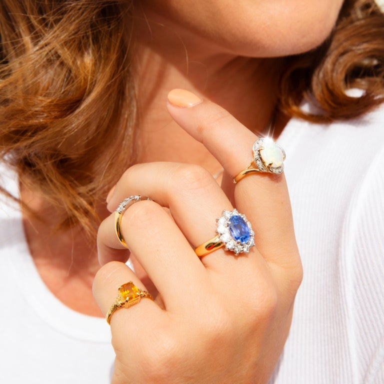 Lovingly crafted in 18 carat gold, this stunning vintage halo cluster ring, Circa 1970s, is a decadent jewel and full of nostalgia. A gorgeous 4.00 carat bright Ceylon-type blue sapphire rests at the centre of a scintillating border of round