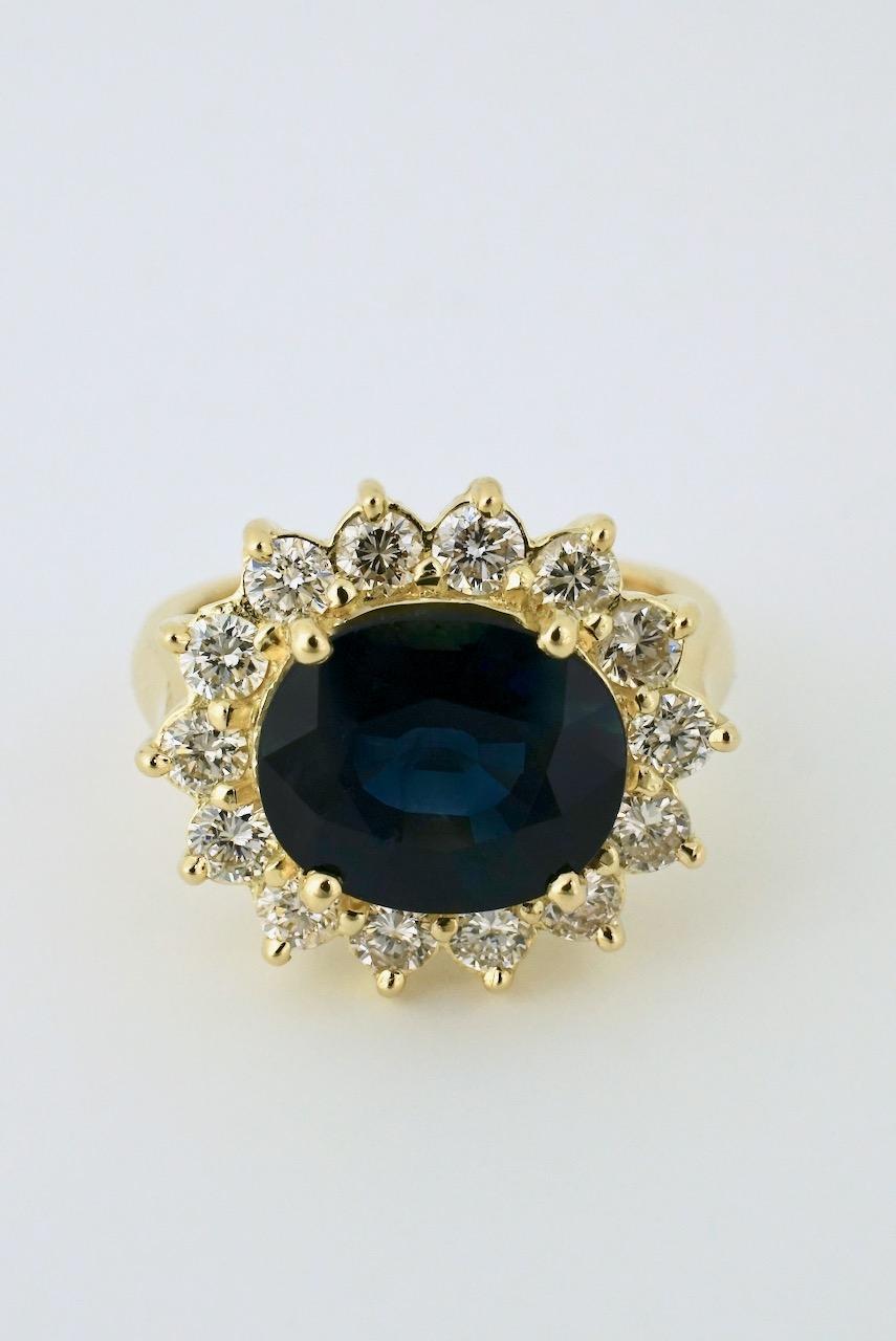 A vintage cluster ring featuring an oval Australian type dark blue sapphire with very good clarity four claw set across the finger and surrounded by a halo of 14 brilliant cut diamonds set in 18k yellow gold on a half dome shank with upswept