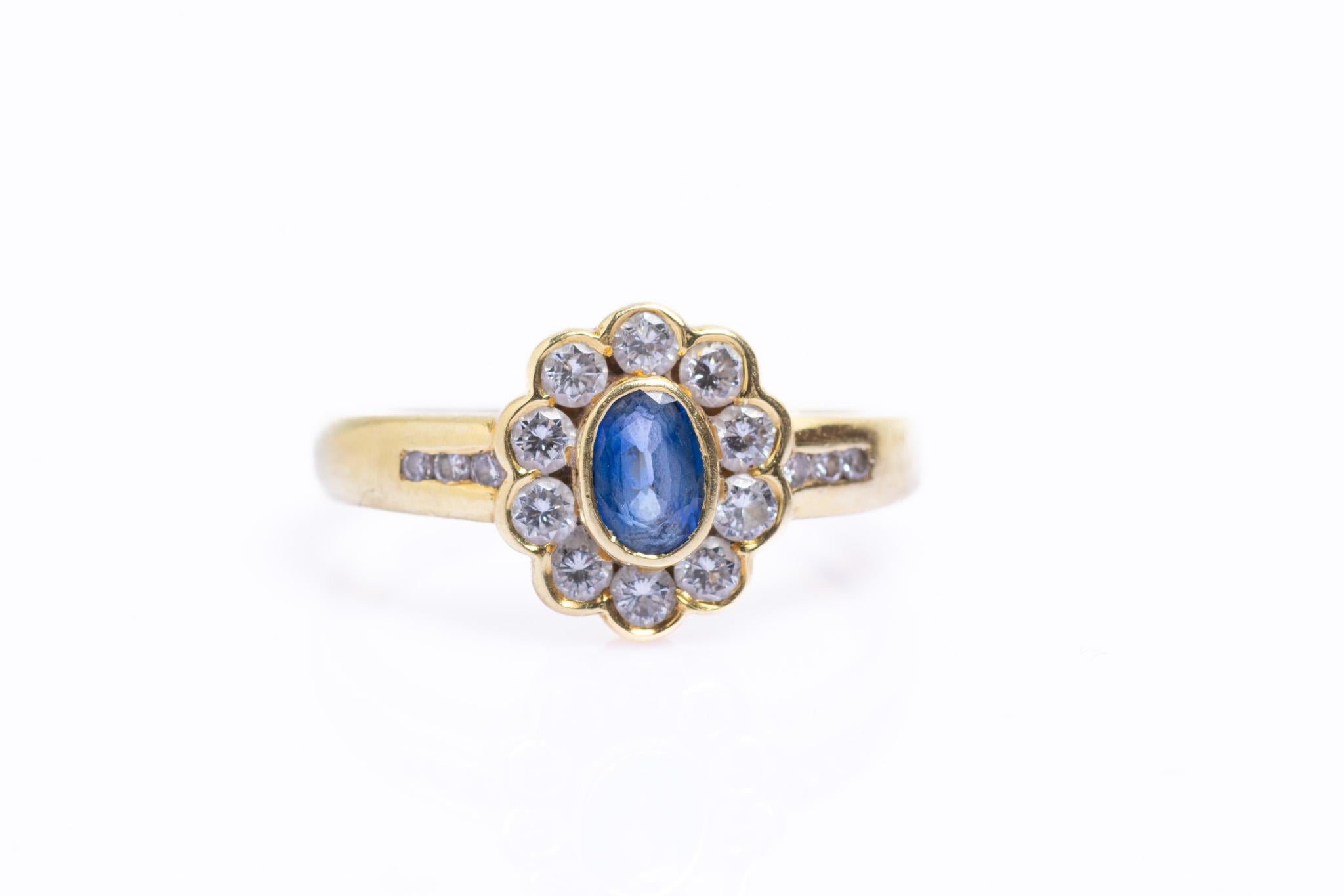 Ein exquisiter Vintage-Ring aus strahlendem 18-karätigem Gelbgold mit einem lebhaften Saphir im Ovalschliff in der Mitte, umgeben von 10 funkelnden Diamanten im Brillantschliff in einem floralen Halo-Design. Die Schultern des Armbands sind mit je