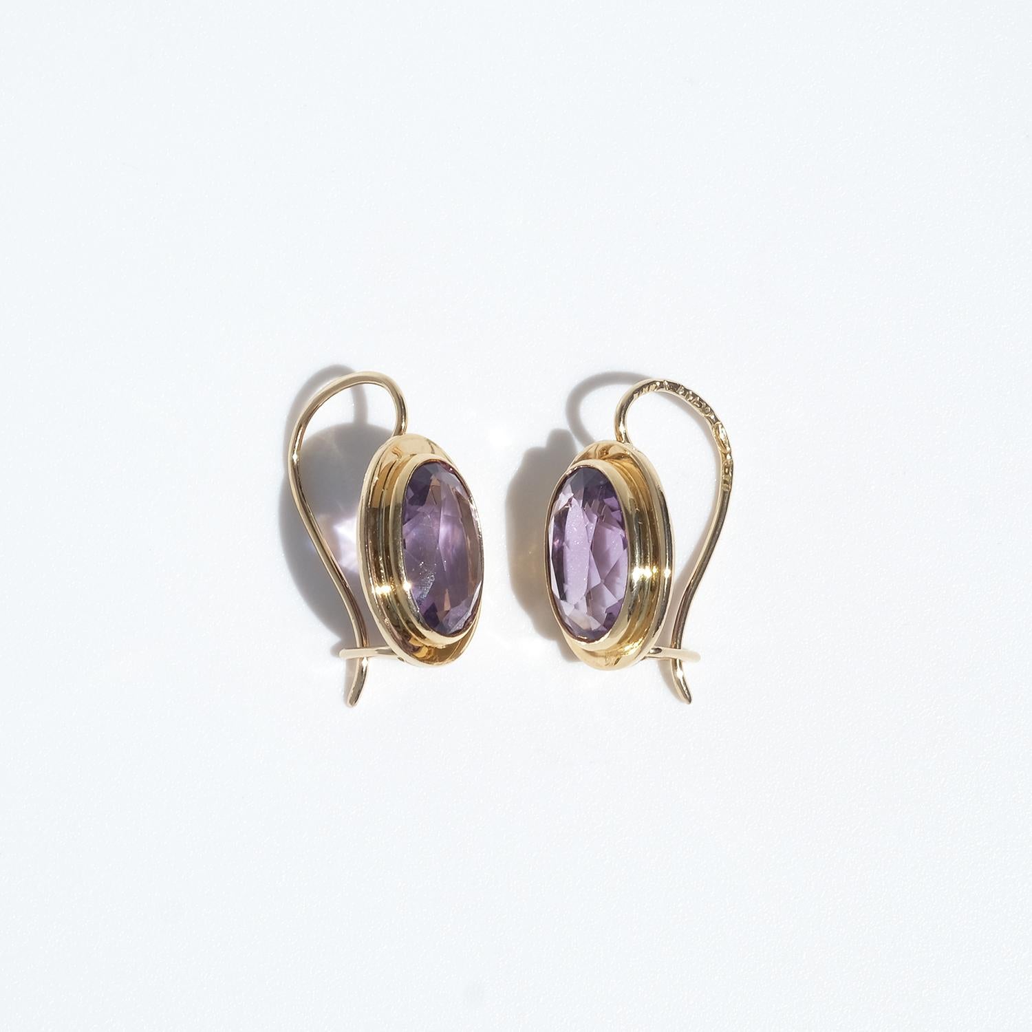 These vintage earrings are crafted in 18 karat gold and feature faceted oval amethysts in a rich, medium purple hue. The gemstones are bezel-set in smooth, slightly raised oval frames that give the earrings depth. They fasten with curved ear wires
