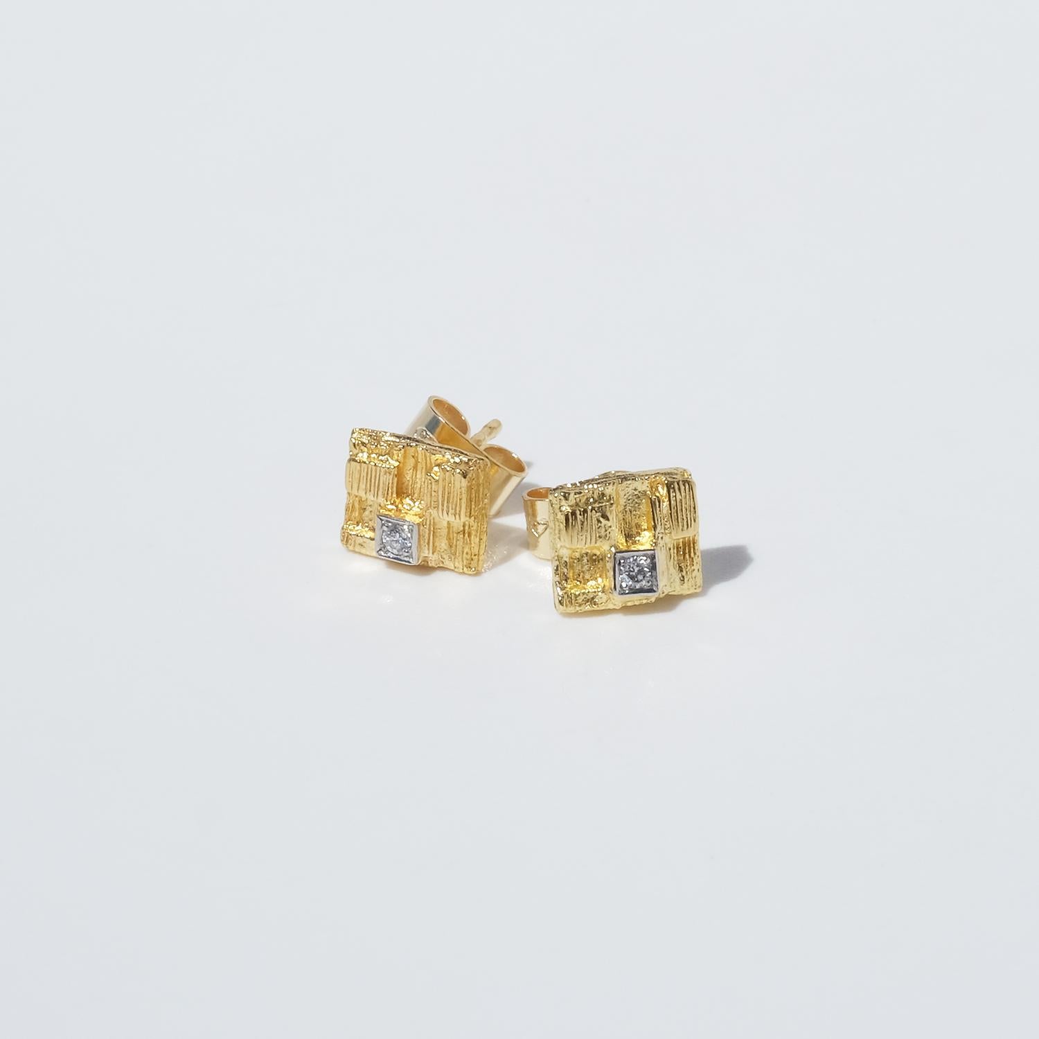 These 18 karat gold stud earrings from Björn Weckström’s Thai Collection feature a rectangular form with a richly textured surface reminiscent of natural, organic structures. A single small diamond is set in the center of each earring, catching the