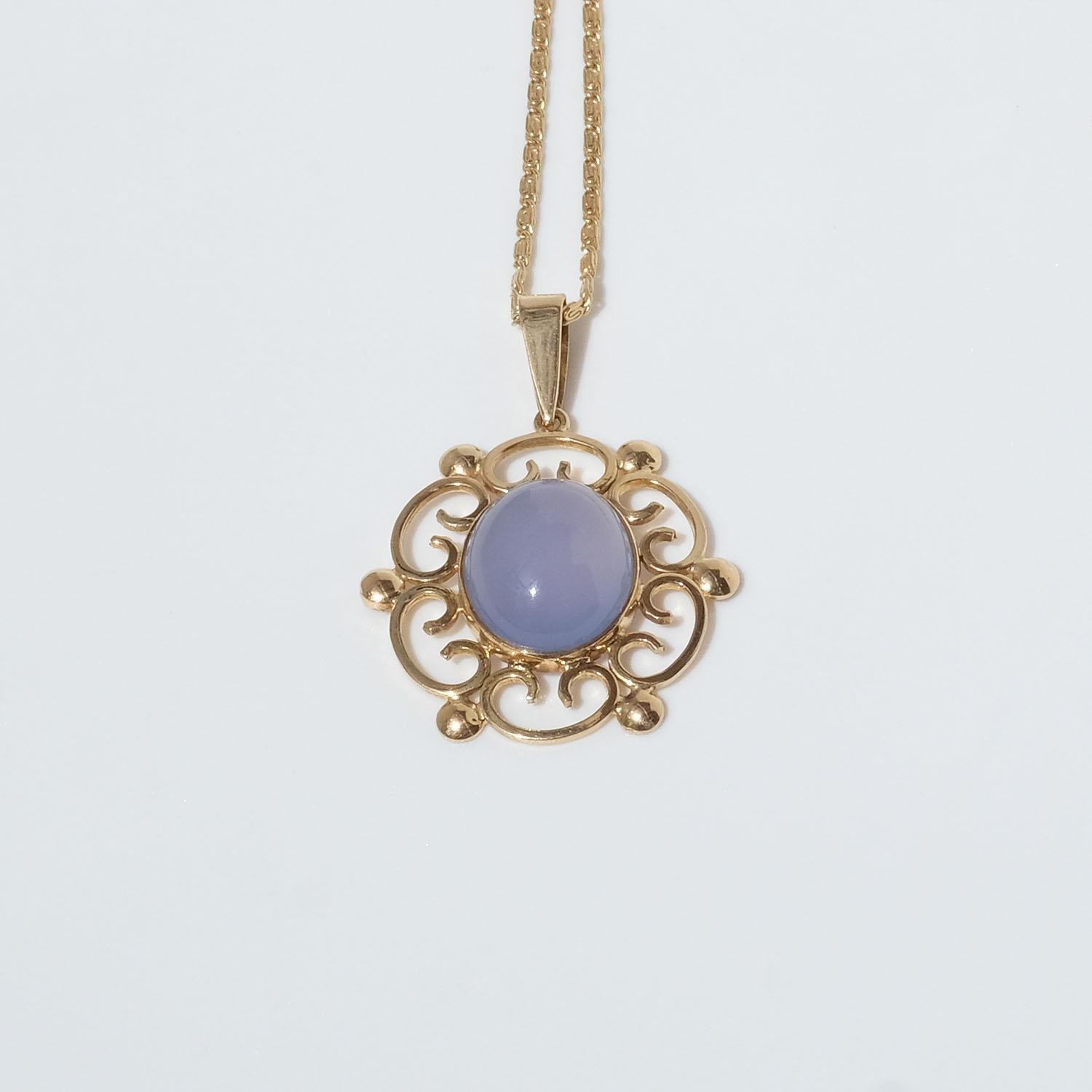 This 18 karat gold pendant features a smooth, oval moonstone with a cabochon cut. The moonstone exhibits a soft, translucent blue hue with a subtle glow that appears to shift and change when it catches the light. Surrounding the moonstone is an