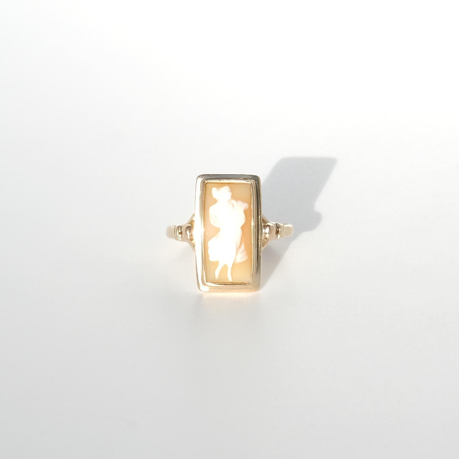 This vintage ring in 18 karat gold features a rectangular bezel setting holding a shell cameo. The cameo depicts a delicate silhouette of a woman with a graceful pose, her profile and flowing dress carved in creamy white against a soft peach