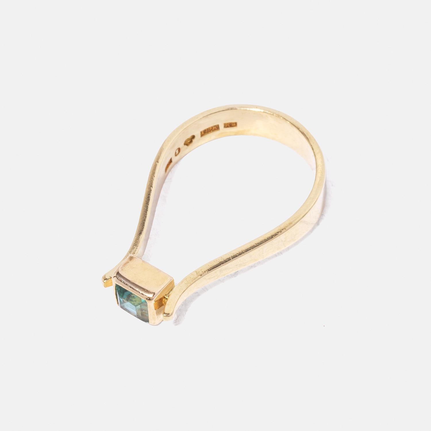 This is an 18-karat gold ring featuring a square green tourmaline. The band has an unusual shape, almost like a loop, giving it a unique and modern appearance. The setting for the tourmaline is simple yet elegant, enhancing the stone's vibrant