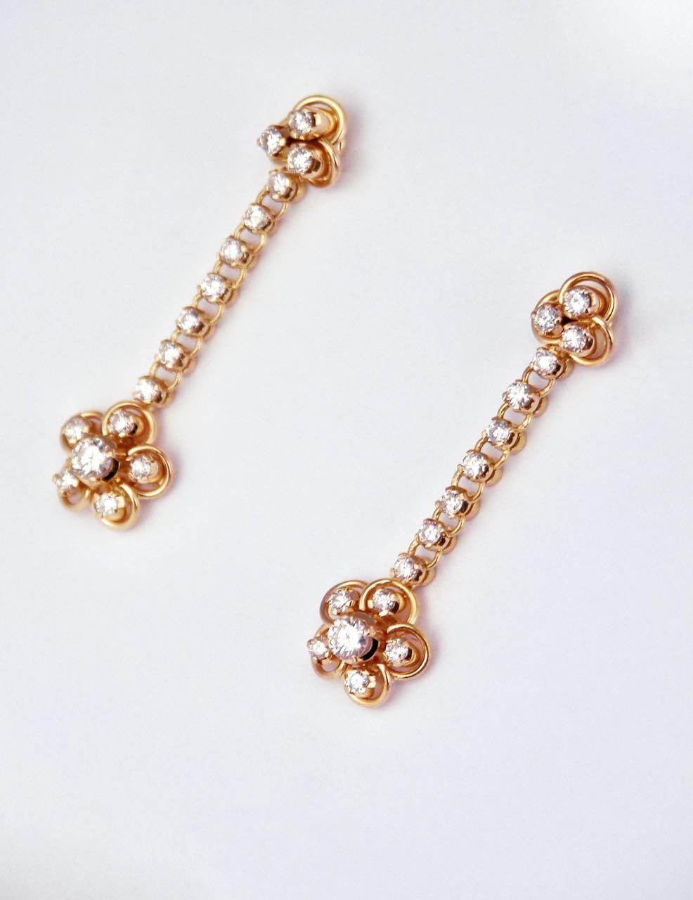 A pretty pair of 18k gold diamond drop earrings; each earring consisting of a delicate cluster design of six brilliant cut diamonds arranged in a flower type motif suspended on an articulated chain set with seven diamonds and below a trefoil shape