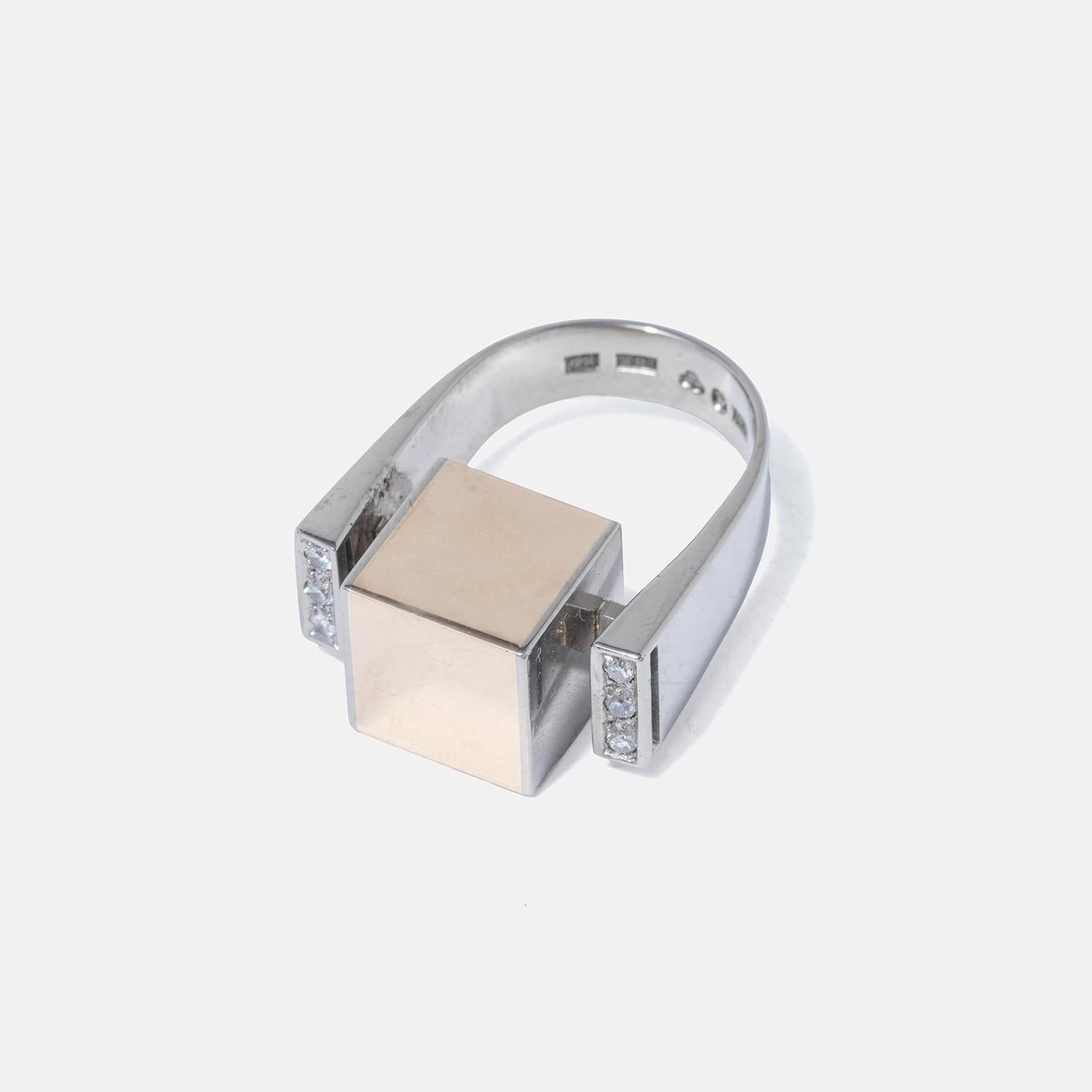 This ring made by the Swedish master Rey Urban showcases a modern design with a wide band that tapers from smaller to wider, crafted from white gold and featuring a prominent, flat square adornment made of polished red gold. Diamonds embellish the