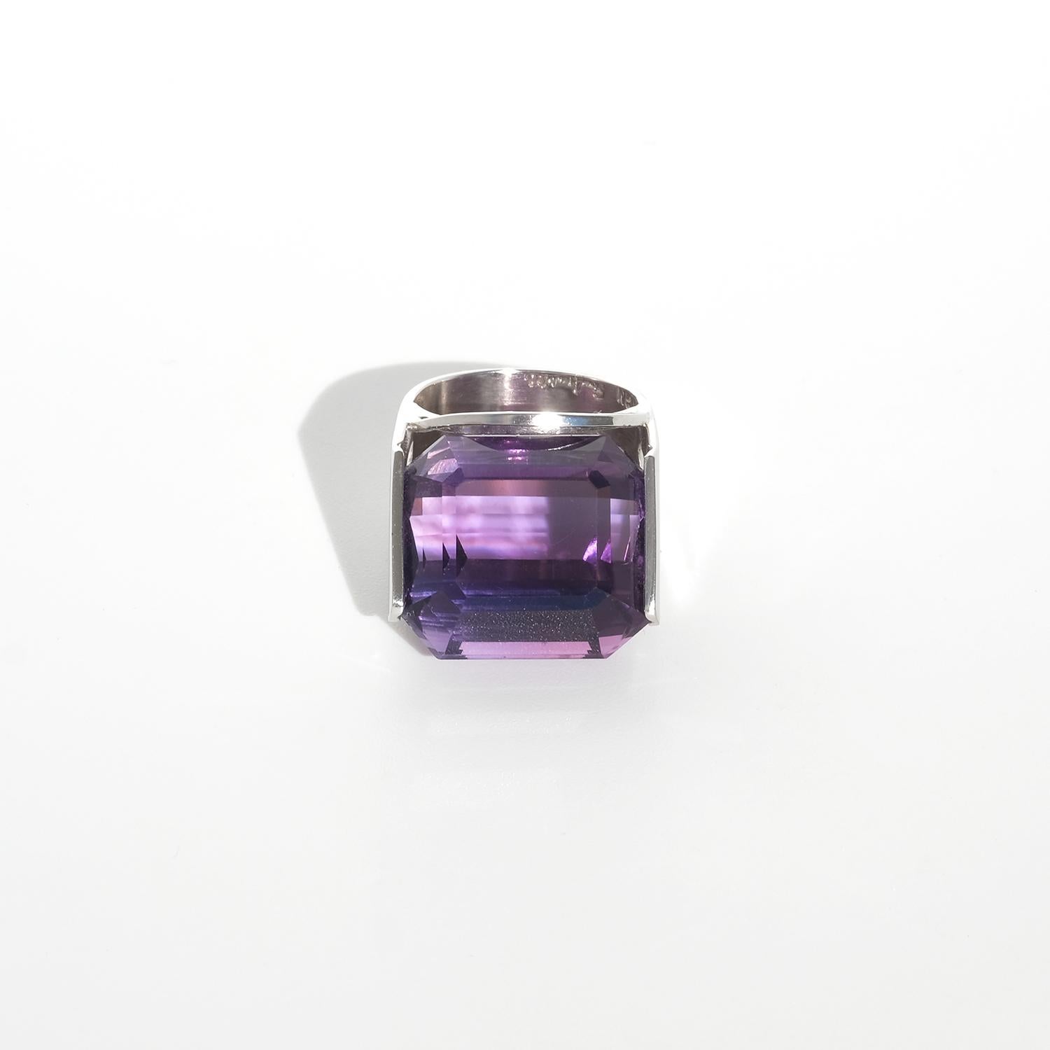 This vintage ring, crafted in white gold, showcases a striking rectangular-cut amethyst with sharp, clean facets that highlight its vibrant and deep violet hue. The stone is securely set in a geometric open bezel, allowing light to illuminate its