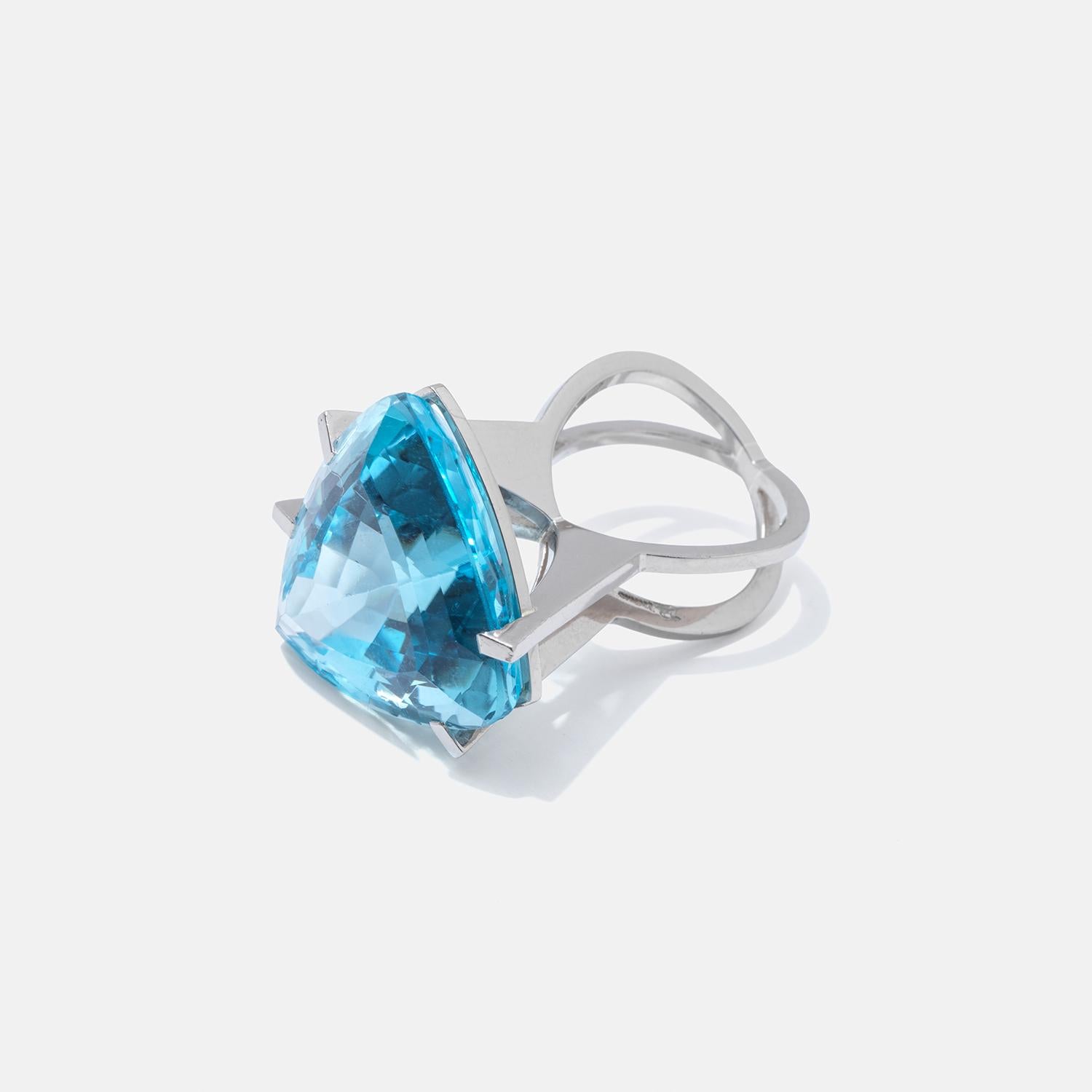 This 18 karat white gold ring features a prominently large blue topaz, boasting an intense shade of azure that catches the eye. The gemstone is cut in a triangular shape, adding a unique geometric appeal to the piece. Its substantial size and bold