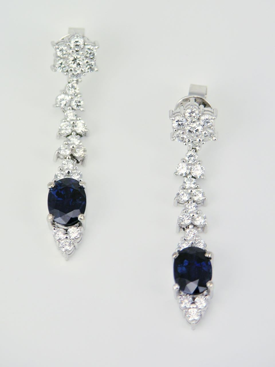 A pair of 18k white gold blue sapphire and diamond drop earrings - each elegant earring consists of an oval dark blue sapphire set in amongst 22 diamonds arranged in leaf shape motifs and suspended below a flower shape cluster with post fittings for