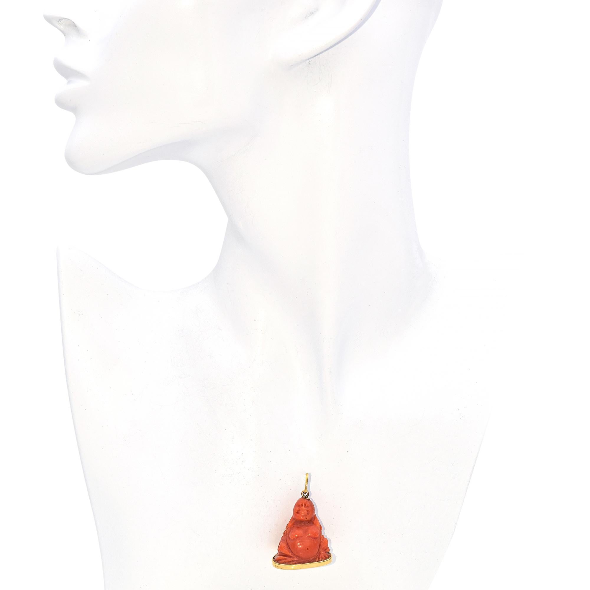 Umarmen Sie die Schönheit der Gelassenheit und des Wohlstands mit diesem exquisiten, handgeschnitzten Buddha-Anhänger aus Koralle, gefasst in 18 Karat Gold. Die warmen, satten Farbtöne der natürlichen Koralle unterstreichen die komplizierten Details