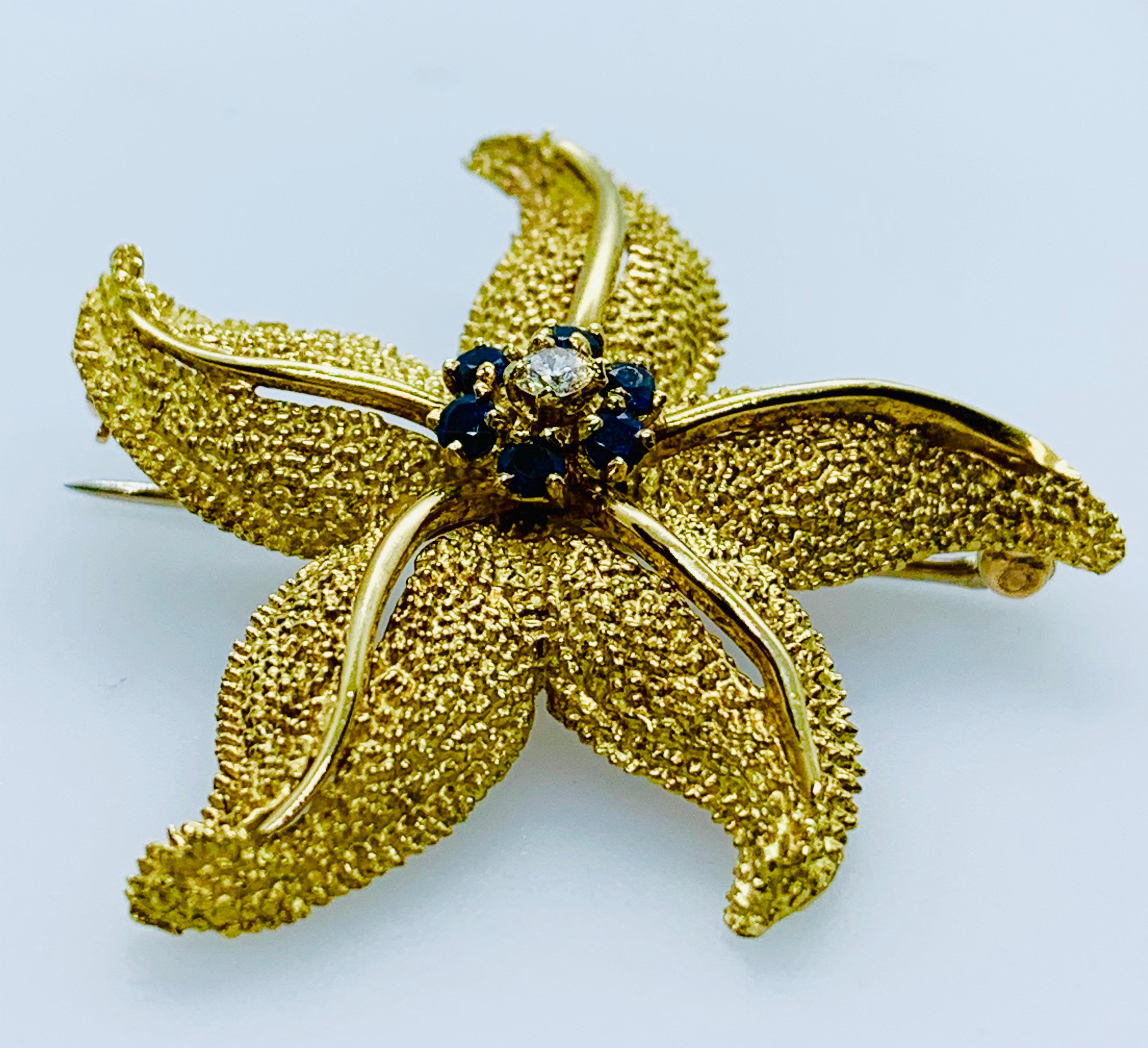 Beautiful Vintage Starfish Brooch! It is made in 18K yellow Gold with a granulated finish mimicking the starfish's texture. At the center of the brooch is a 0.08 carat Brilliant Diamond that is surrounded by 6 round blue sapphires. The brooch