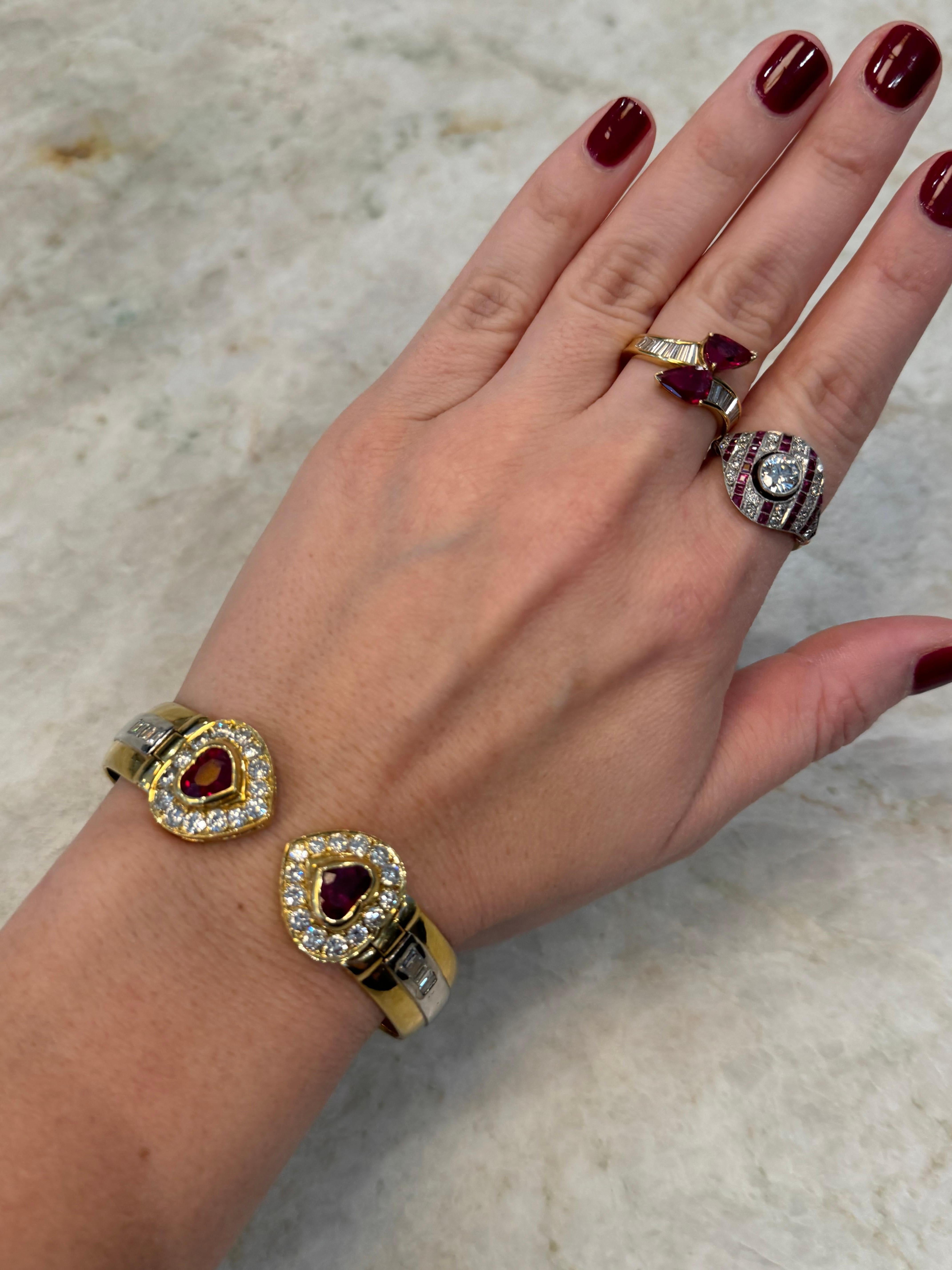 An eye-catching vintage bangle bracelet crafted in 18K yellow gold, featuring two heart-shaped rubies at the center, weighing approximately 2.84 carats combined. Each ruby is framed by a halo of round brilliant diamonds, and the shoulders are