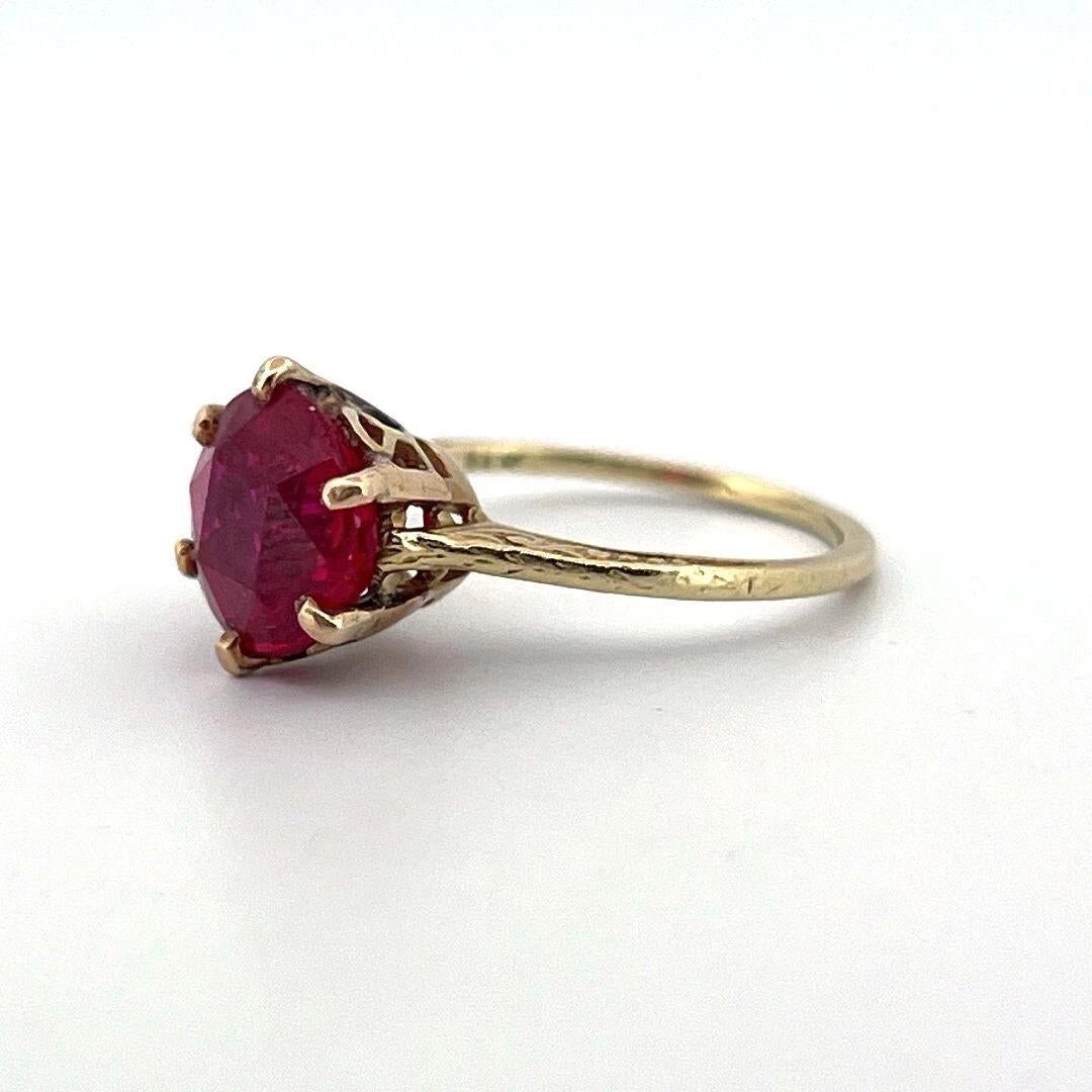 This beautifully detailed vintage ring is crafted in solid 18 karat yellow gold and features a vibrant synthetic ruby, measuring approximately 8.40 mm in diameter. The ruby is prong-set in an ornate openwork crown-style solitaire mounting, with