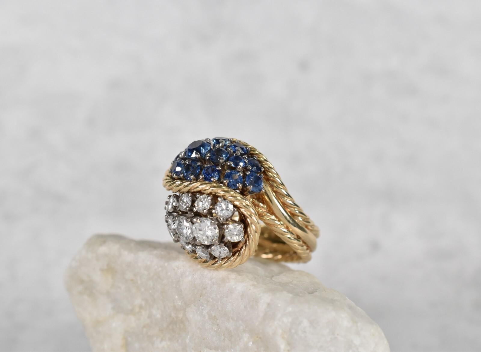 This bold 18k yellow gold cocktail ring features a dramatic bypass design, set with clusters of vibrant blue sapphires and dazzling white diamonds. The twisted gold rope detailing gracefully wraps around each section, adding texture and vintage