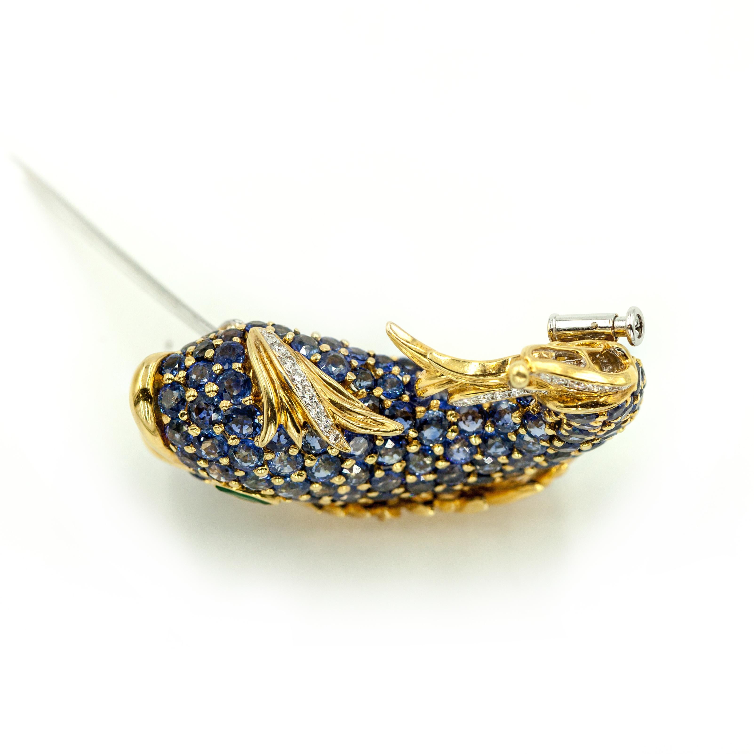 A striking and whimsical 18K yellow gold diamond sapphire fish brooch. The body is pave set round cut nature sapphires in vivid blue creating a rich scaled texture. A marquise cut emerald is used for the eye, the tail and fin accents are embellished