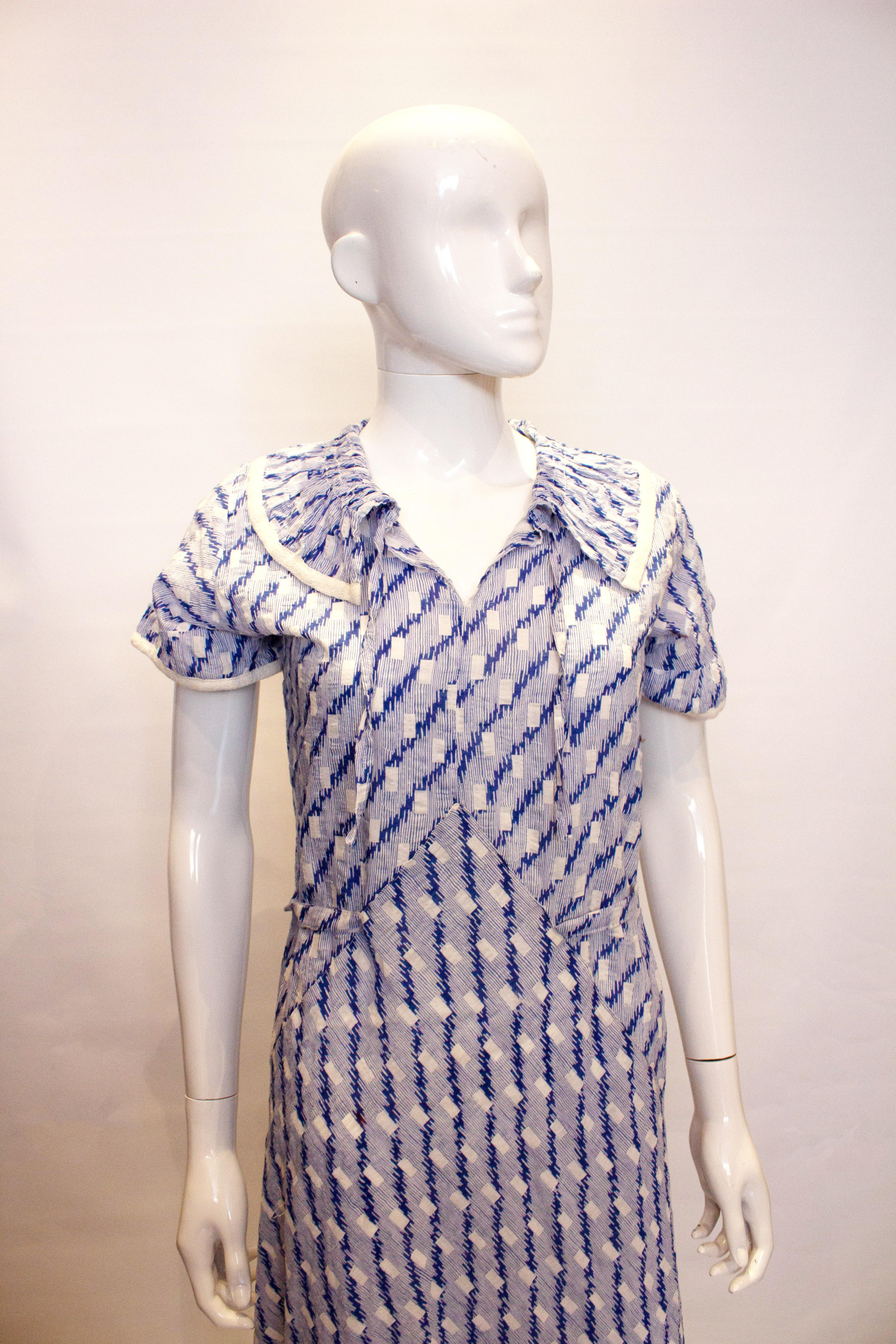 A pretty and easy to wear vintage cotton day dress from the 1920s.  In a blue and white print, the dress has a gathered collar with white trim and tie at the neck. It has a tie back at waist leval and white trim on the edge of the sleaves.