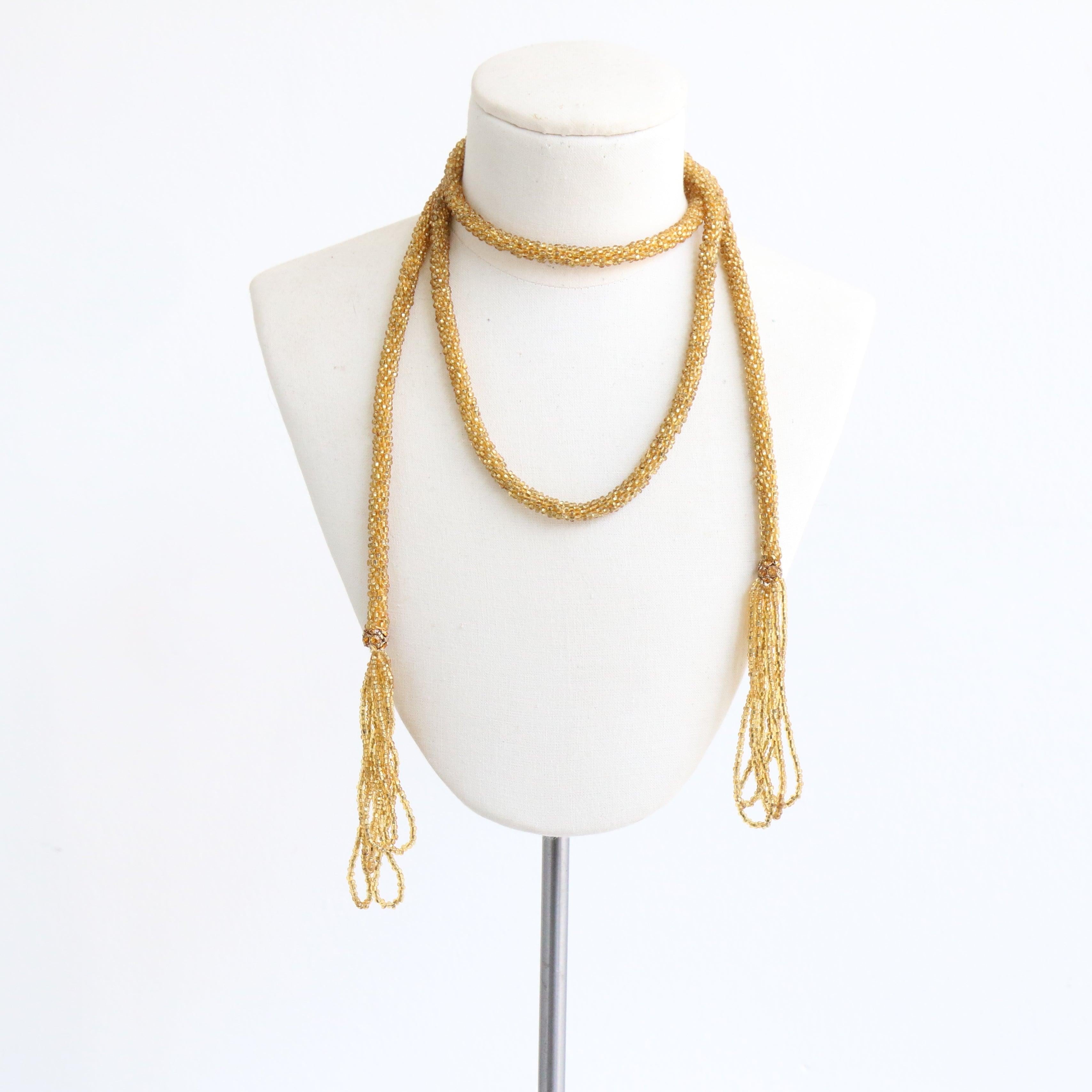 Ce magnifique collier lariat en perles de verre en or jaune des années 1920 est une pièce rare.

Son tube de perles tressées est terminé à chaque extrémité par des détails de pompons décoratifs bouclés tombant de perles de strass jaunes serties en