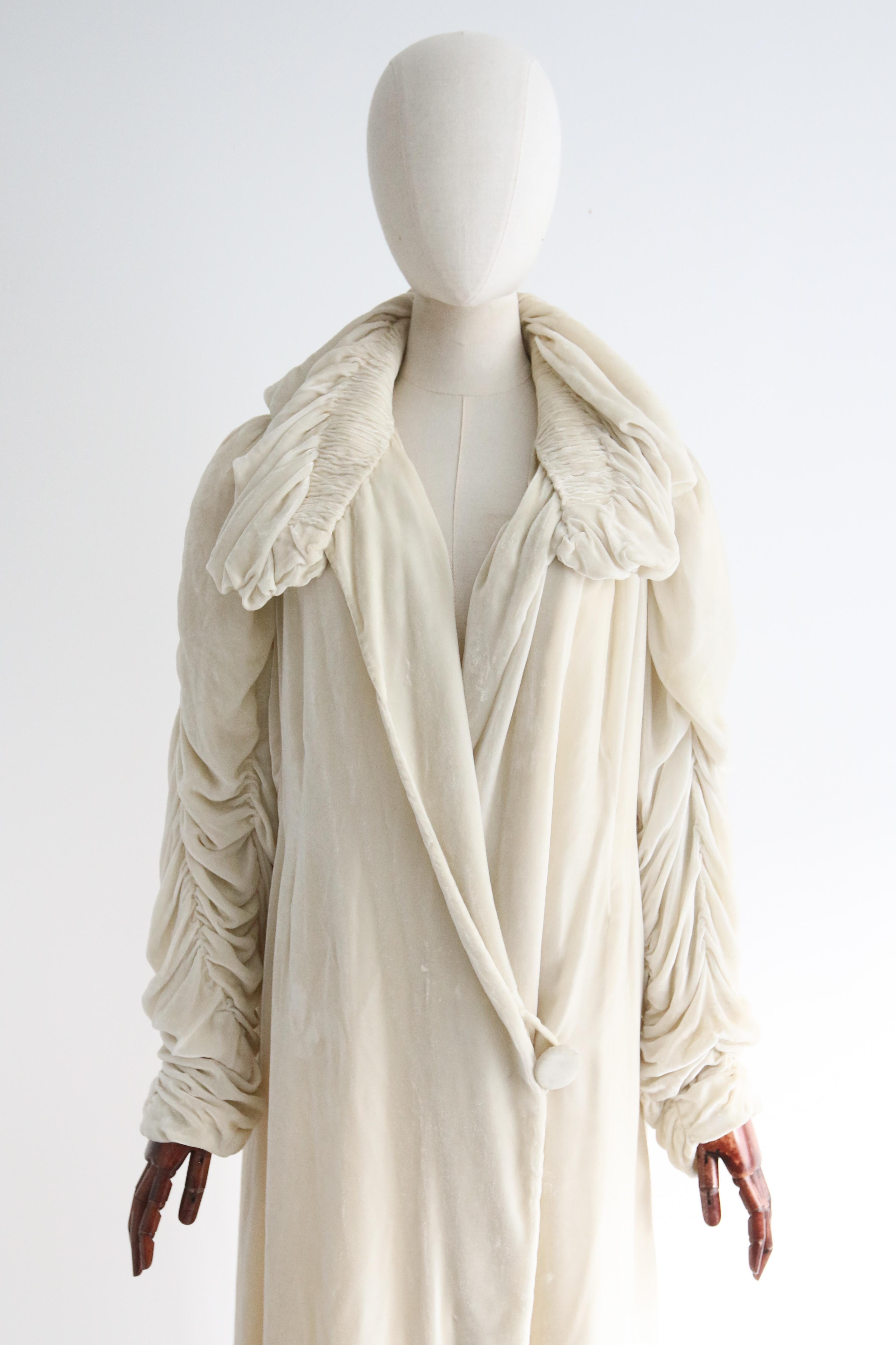 This truly majestic 1920's silk coat, rendered in the most sumptuous shade of ivory silk velvet and lined in the softest silk, is a rare piece to behold.
The deep V shaped neckline of the coat is framed by a pleated and gathered ruffled collar,