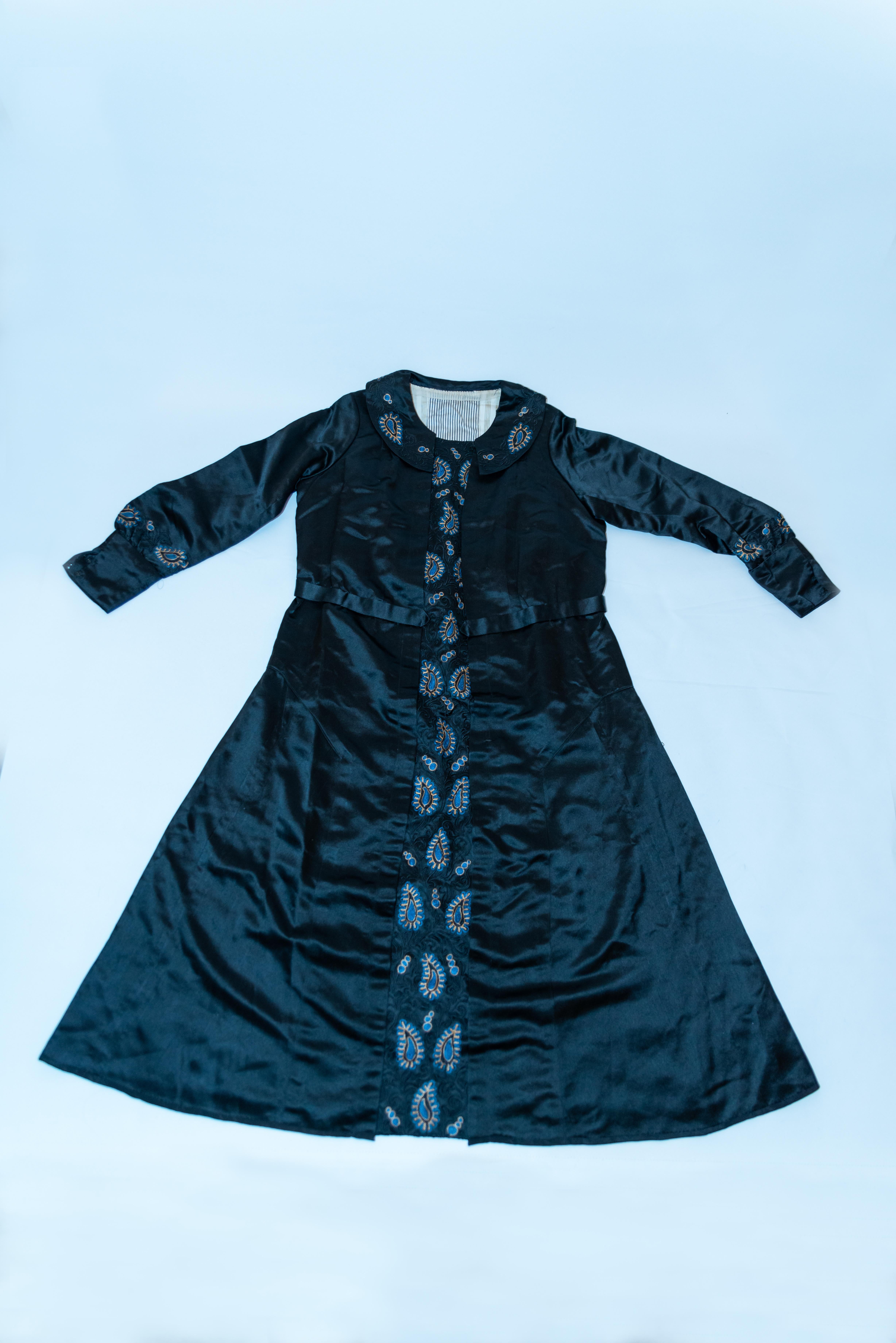 Ein schickes schwarzes Satinkleid aus den 1920er Jahren. Das Mantelkleid ist gefüttert und hat blaue und goldene Verzierungen am Kragen, an den Manschetten und am Mittelteil.

Maße: Büste bis zu 39'', Länge 45''