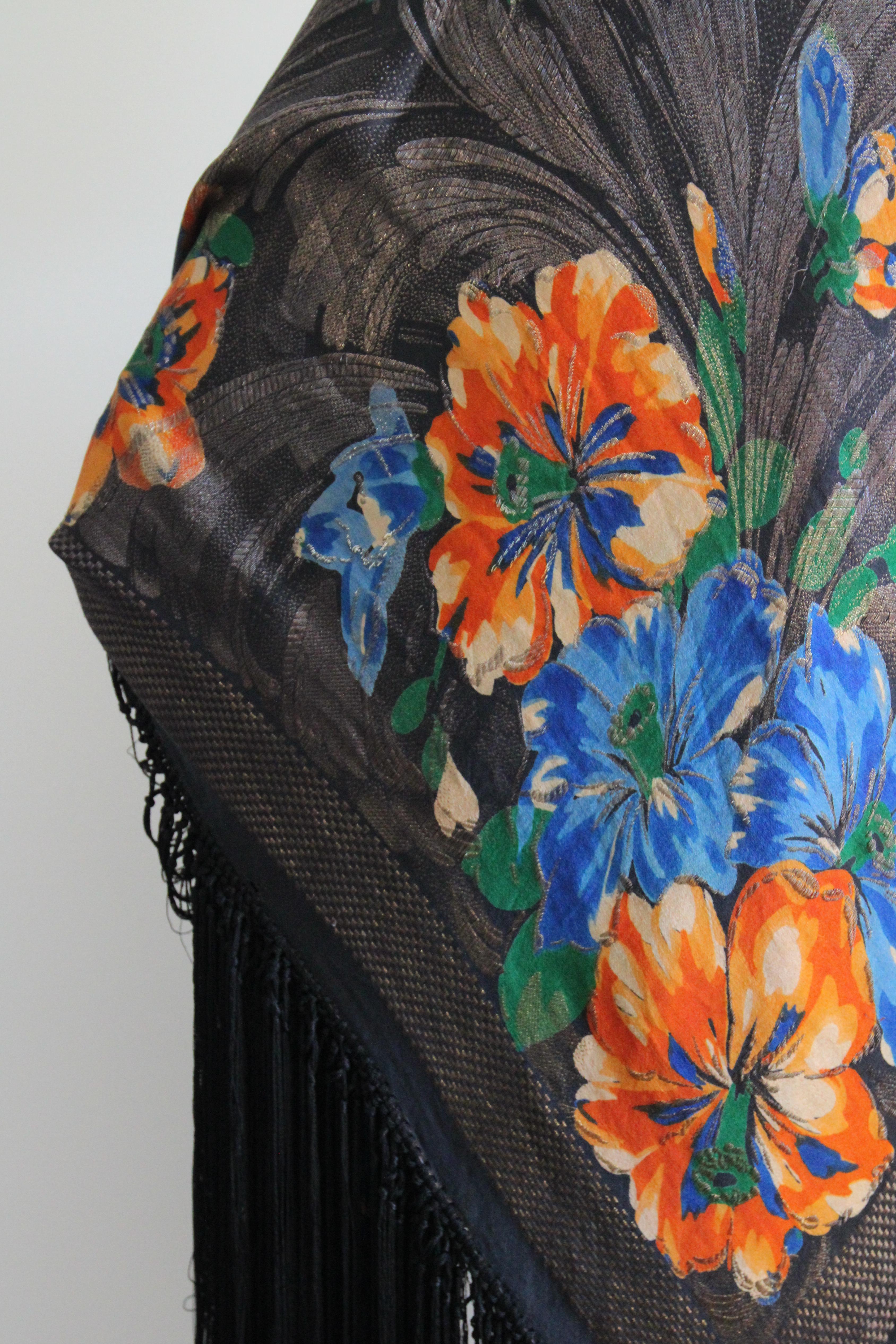 Das atemberaubende Seidenlamé mit dem auffälligen Blumenmuster in Orange- und Blautönen mit grünen Akzenten auf einem tiefschwarzen Untergrund und den blauen und goldenen Lamé-Fäden ist genau das Richtige, um Ihrer Garderobe für die Saison einen