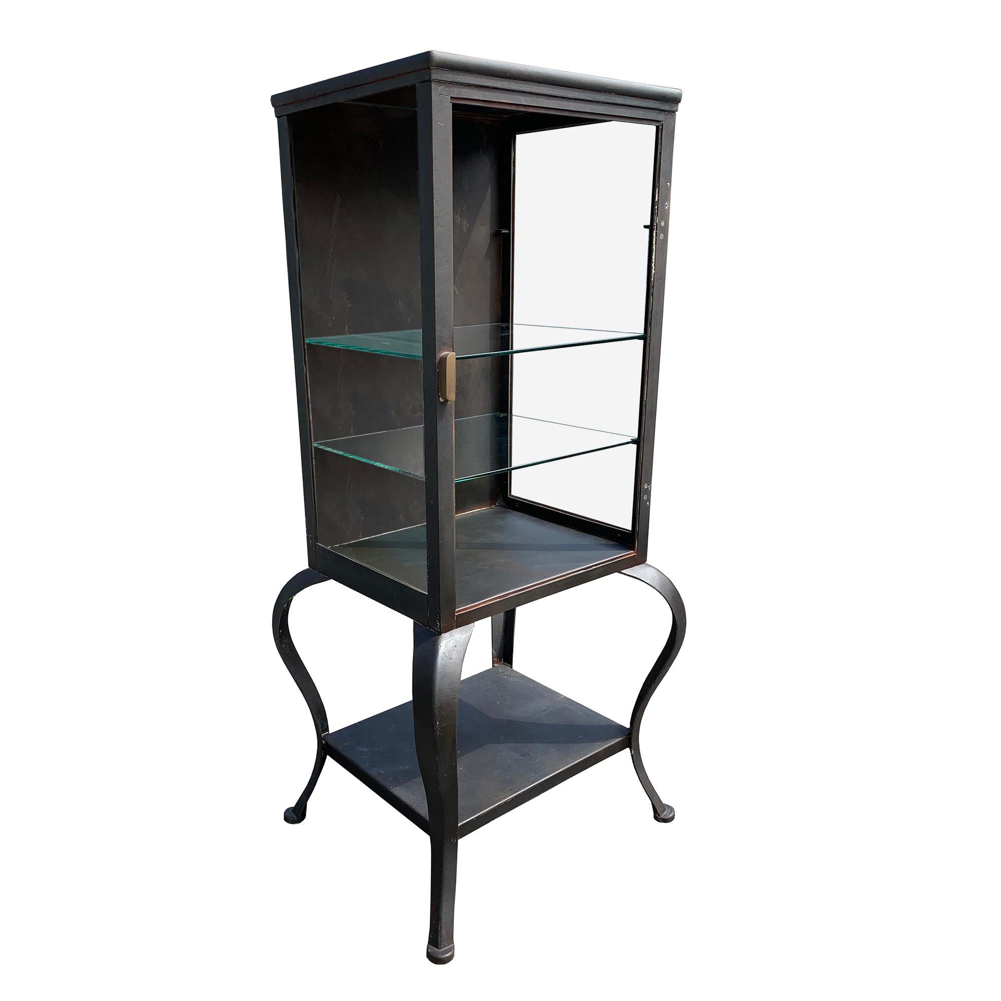 1920s steel medical glass cabinet. This industrial style cabinet includes 2 adjustable glass shelves and a bottom fixed metal shelf. Good rustic condition, this piece is heavy duty steel but is antique and as is, the door was removed at some point