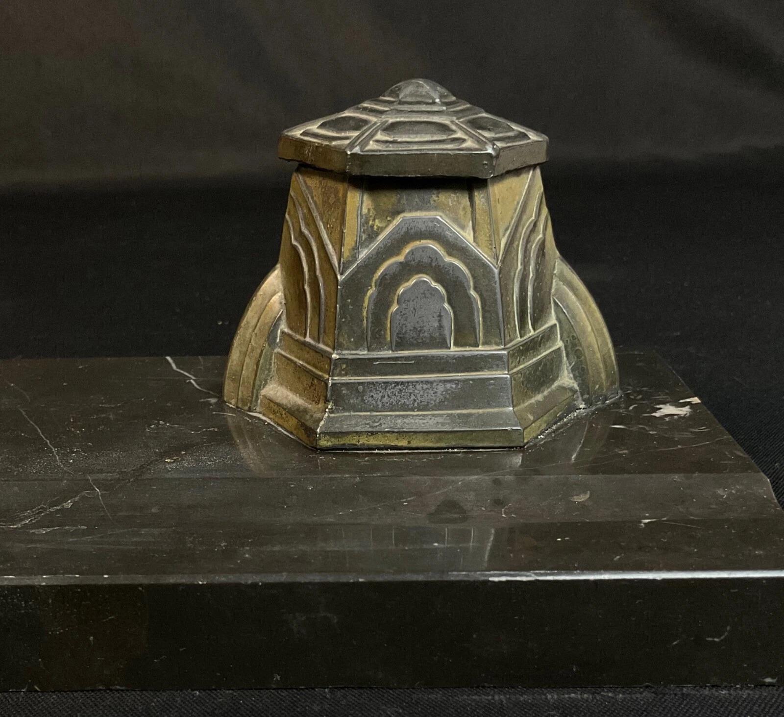 Se trata de un juego de tintero vintage hecho a mano de los años 30, un ejemplo clásico del periodo Art Déco. La pieza presenta un tintero hexagonal de metal con tapa abatible, montado sobre una sólida base de mármol negro. Las líneas limpias, la