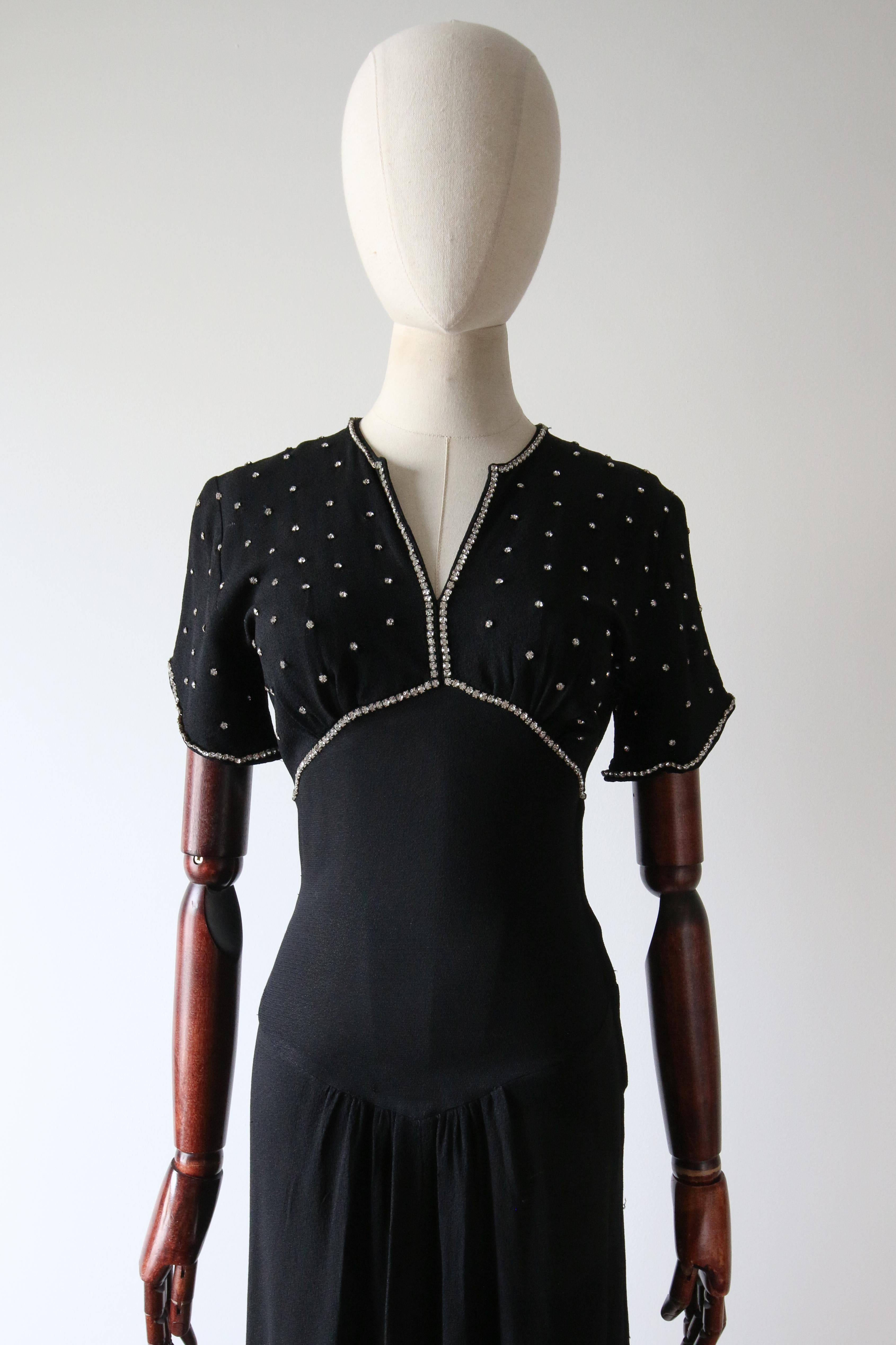 Cette époustouflante robe du soir en crêpe de soie noir des années 1930, décorée de strass argentés sertis, est exactement la pièce qu'il vous faut pour cette soirée spéciale.

Le décolleté en V de la robe est encadré par une bordure de strass